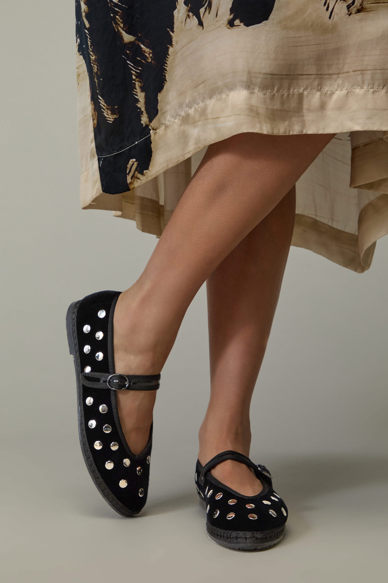 A person stands with legs crossed, wearing a beige and black skirt and Studded Mary Jane Velvet Shoes.