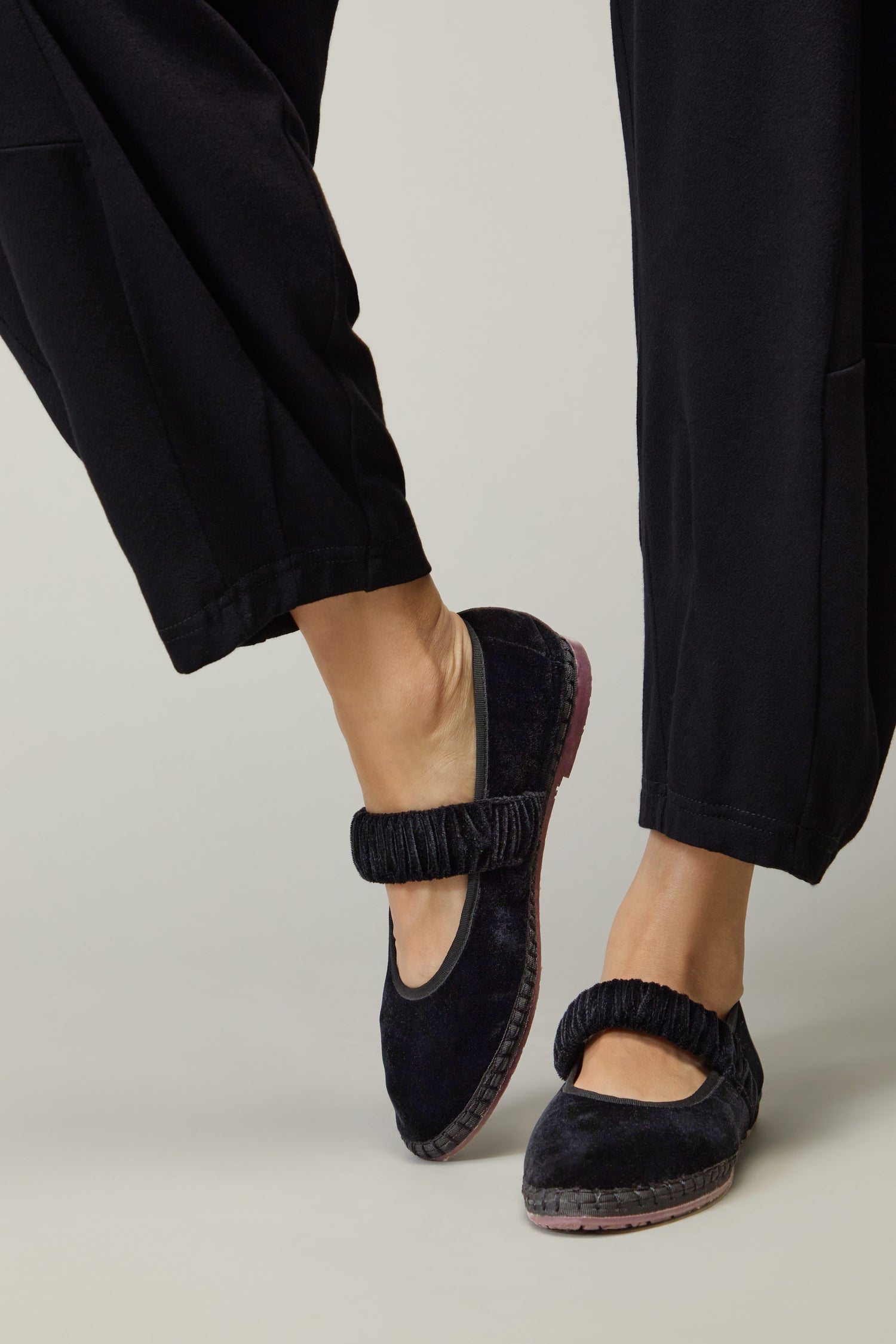 Wearing Mafalda Black Velvet Shoes—handmade in Spain with elastic straps and black velvet, embodying slow fashion—paired with black pants, one foot slightly raised.