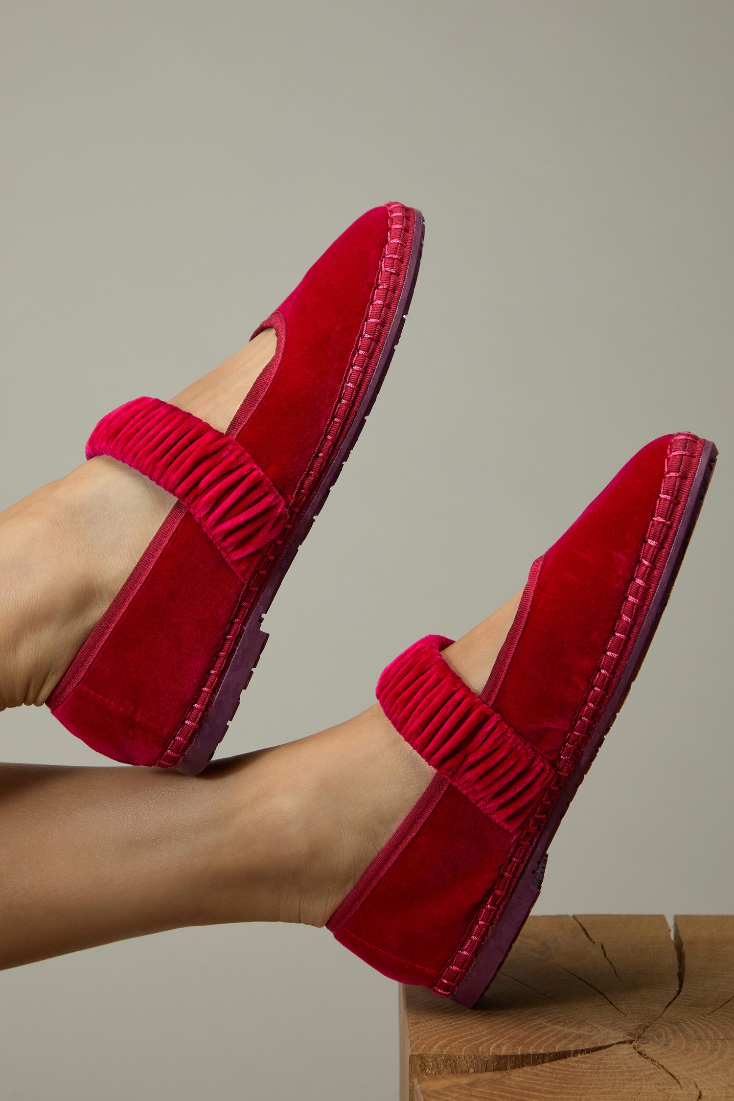 Two feet in Mafalda Fuchsia Velvet Shoes with elastic straps, handmade in Spain with a recycled rubber sole, are shown resting against a neutral background and wooden surface.