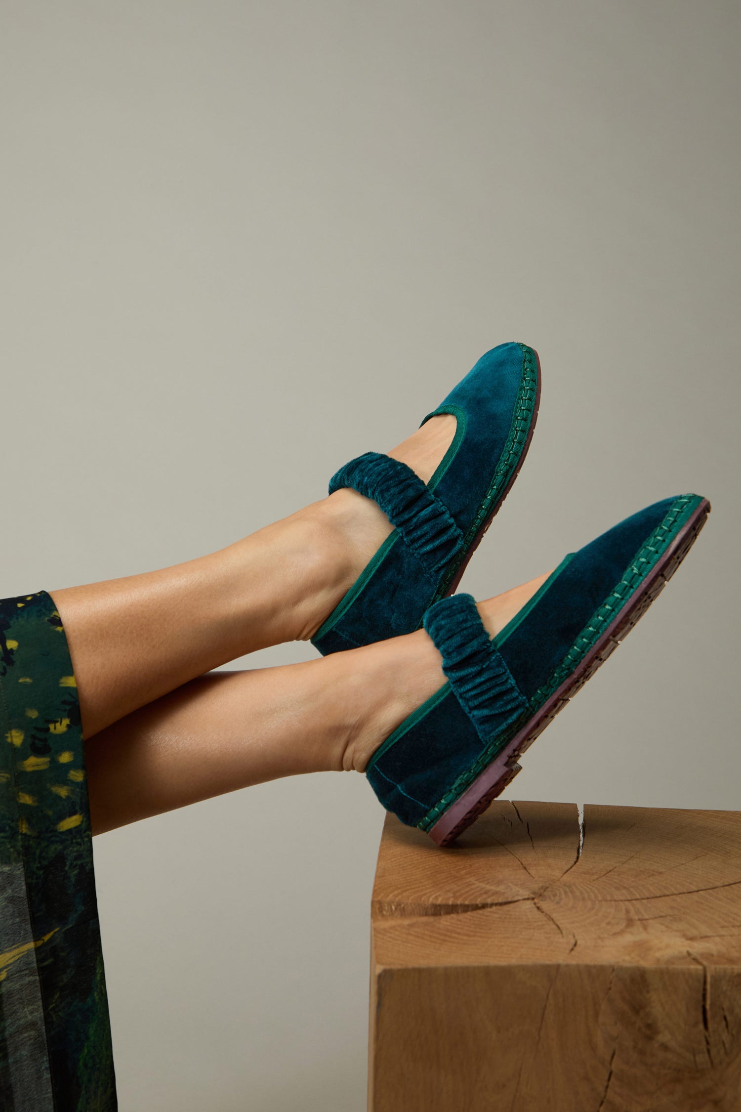 A person wears Mafalda Teal Velvet Shoes—featuring ruched straps and recycled rubber soles—while resting their feet on a wooden surface against a plain background.
