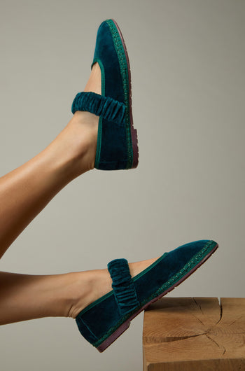 A person with legs raised wears Mafalda Teal Velvet Shoes featuring textured straps and a recycled rubber sole, resting one foot on a wooden surface against a plain background.