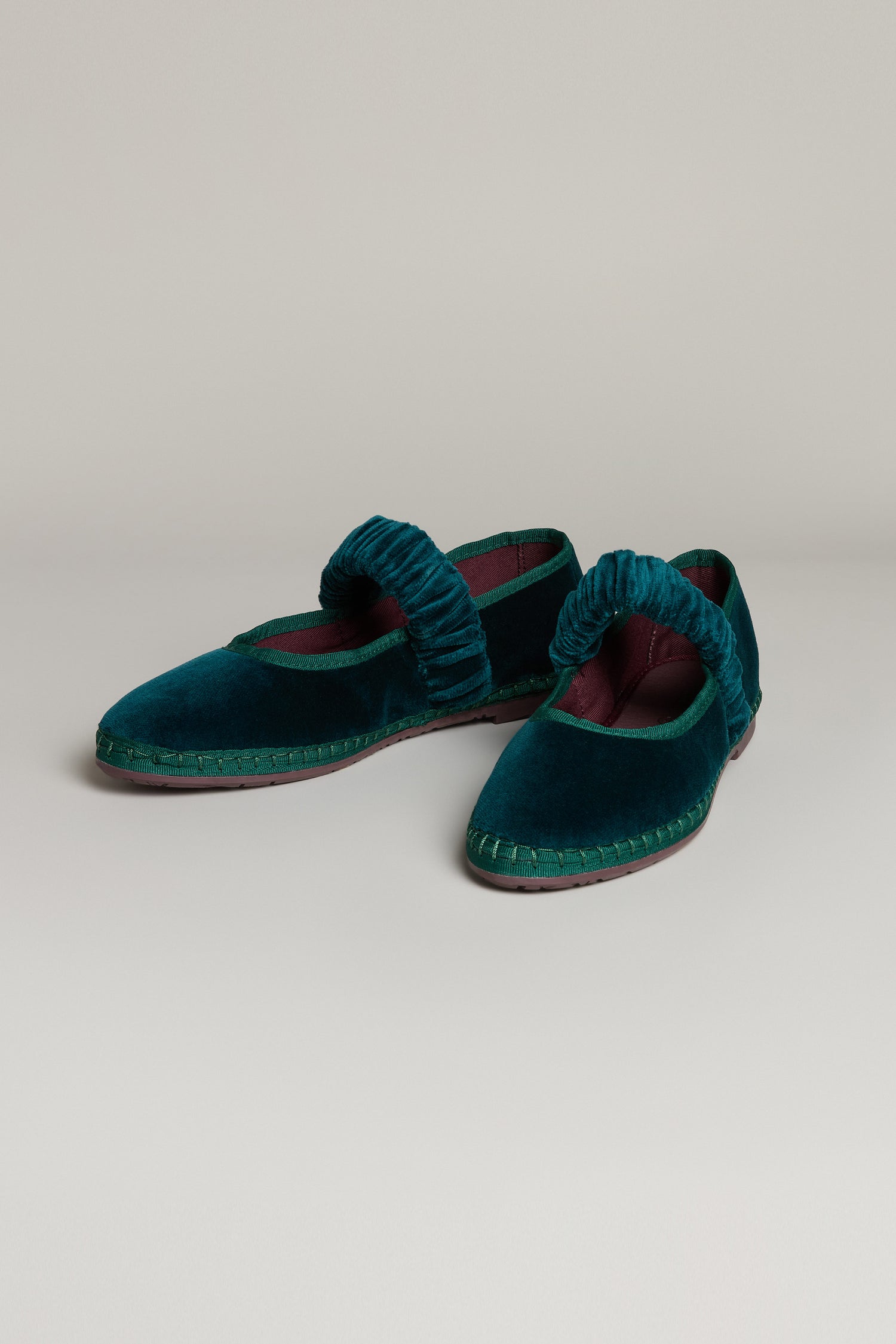 The Mafalda Teal Velvet Shoes feature ruched straps, a dark maroon lining, and a recycled rubber sole. Handmade in Spain from velvet, this flat pair is shown against a neutral background.
