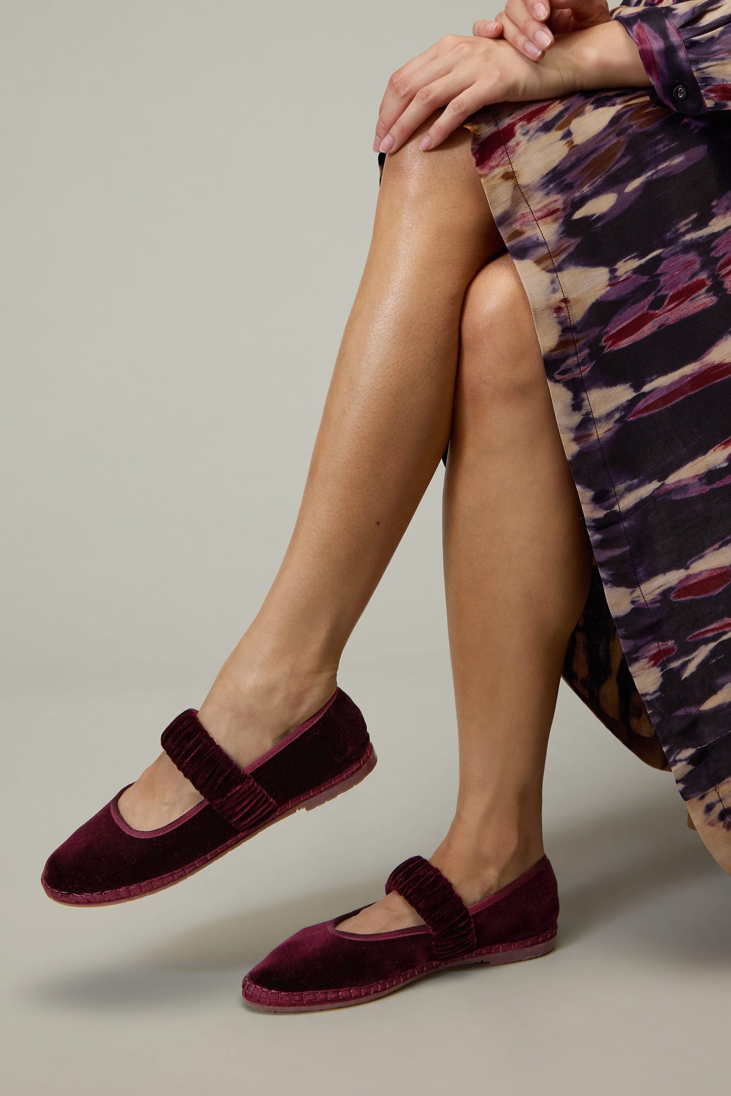 A person in a patterned dress sits with legs crossed, displaying Mafalda Wine Velvet Shoes—crafted with matching straps and an elegant, handcrafted finish inspired by Spanish heritage.
