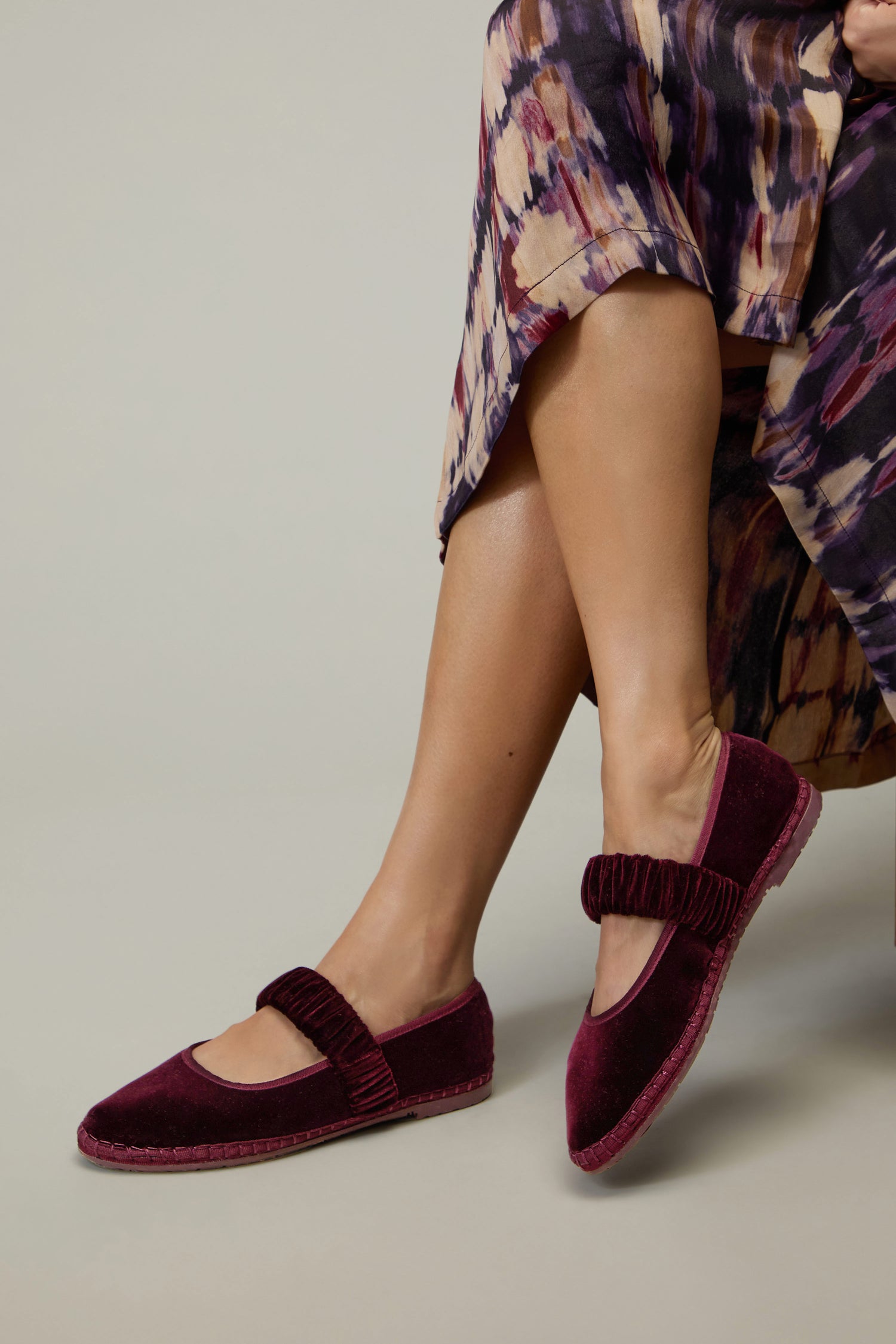 A person wearing a patterned skirt and Mafalda Wine Velvet Shoes handcrafted in Spain with a ruched strap, sitting with legs crossed.
