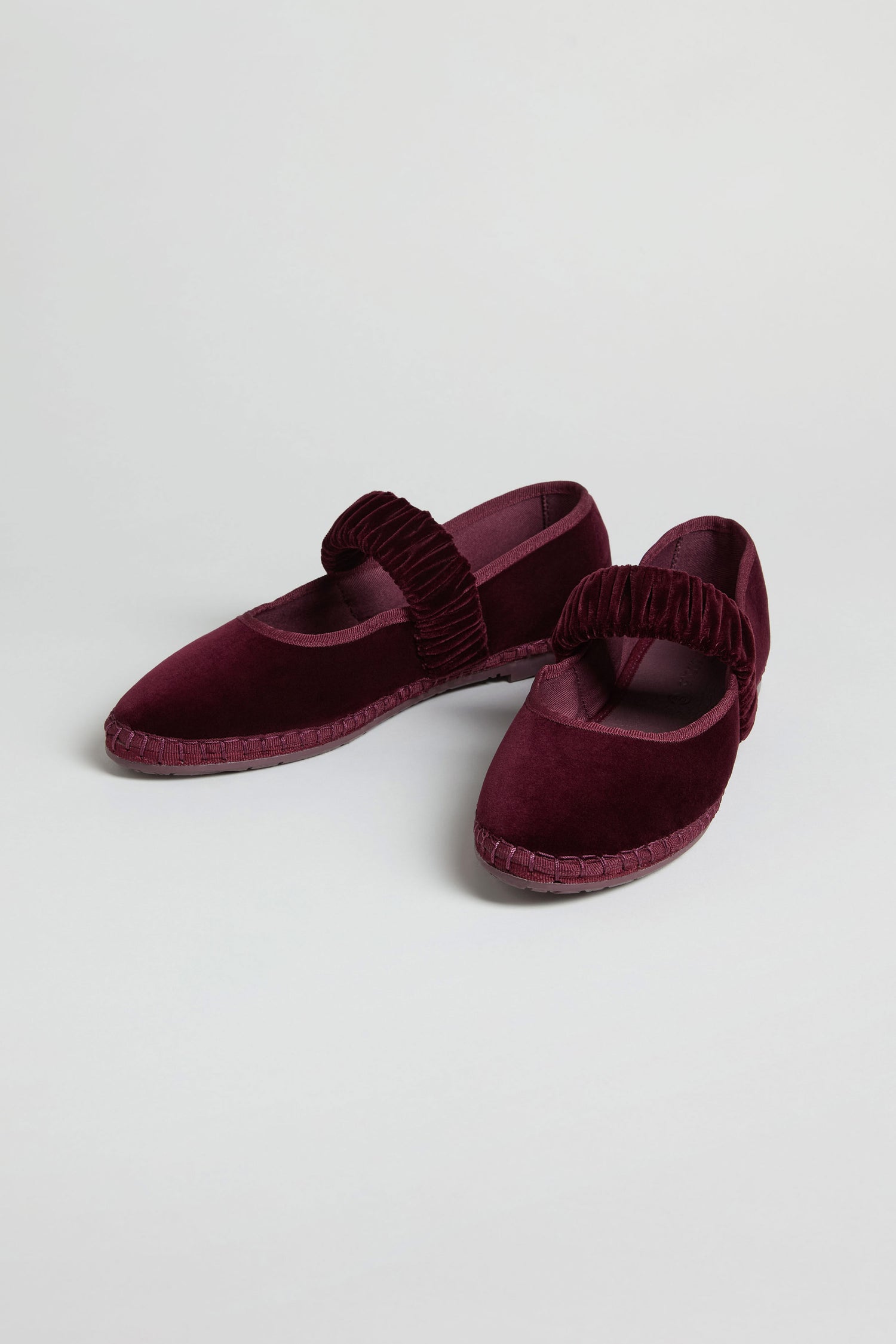 The Mafalda Wine Velvet Shoes, handcrafted in Spain, feature thick ruched straps and flat soles. This maroon Mary Jane pair is displayed on a plain white background.
