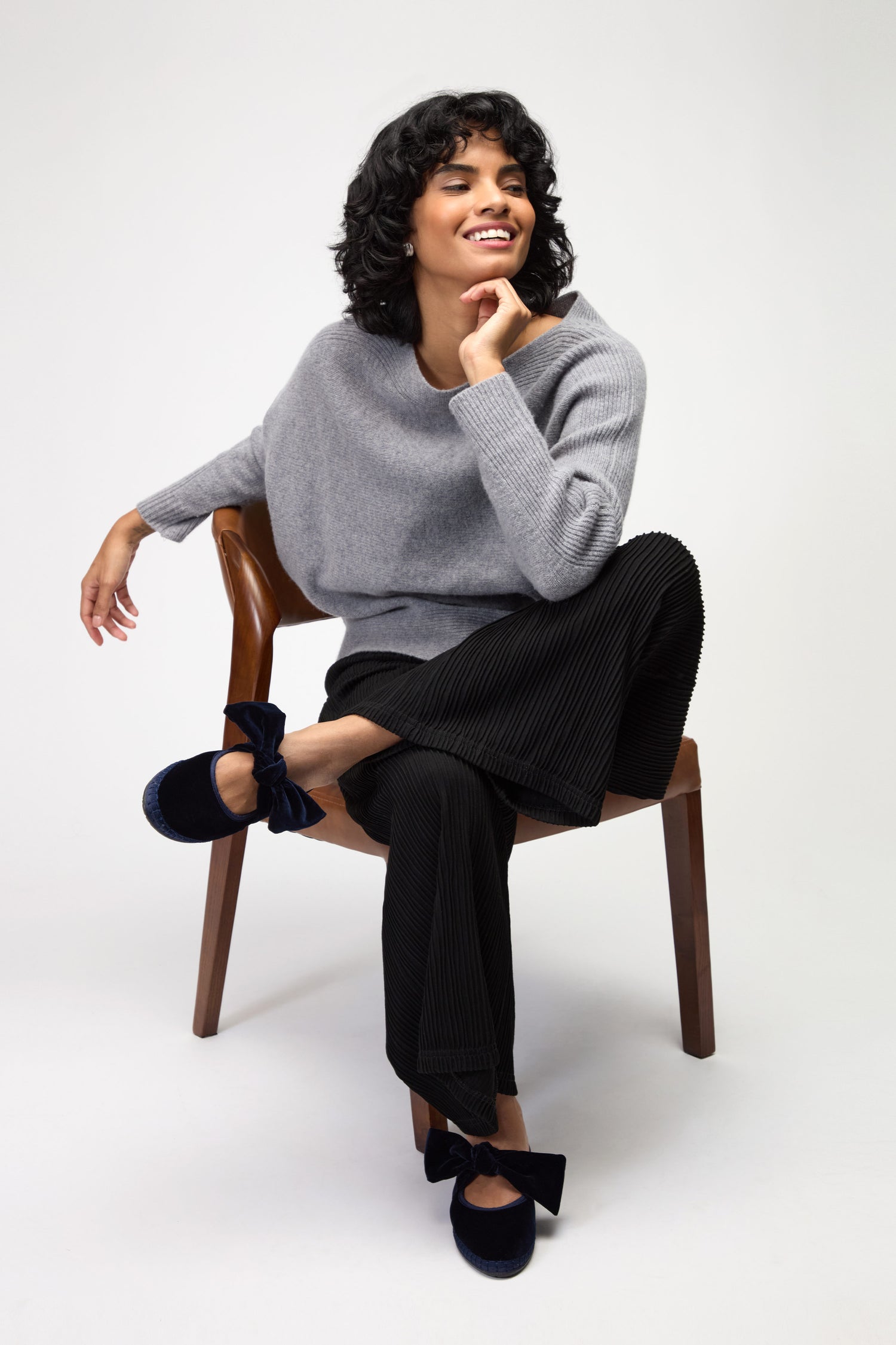 A person with short curly hair sits on a wooden chair, wearing a gray sweater, black pleated pants, and navy Lyra Bow velvet shoes from Flabelus, smiling and resting an arm on the chair back.
