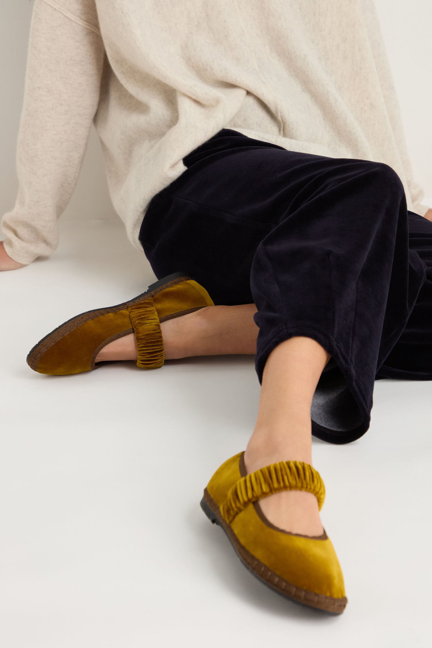 A person sits on the floor wearing black velvet pants, a beige sweater, and Mafalda Ochre Velvet Shoes—mustard yellow velvet footwear featuring ruched straps and a recycled rubber sole, handmade in Spain.