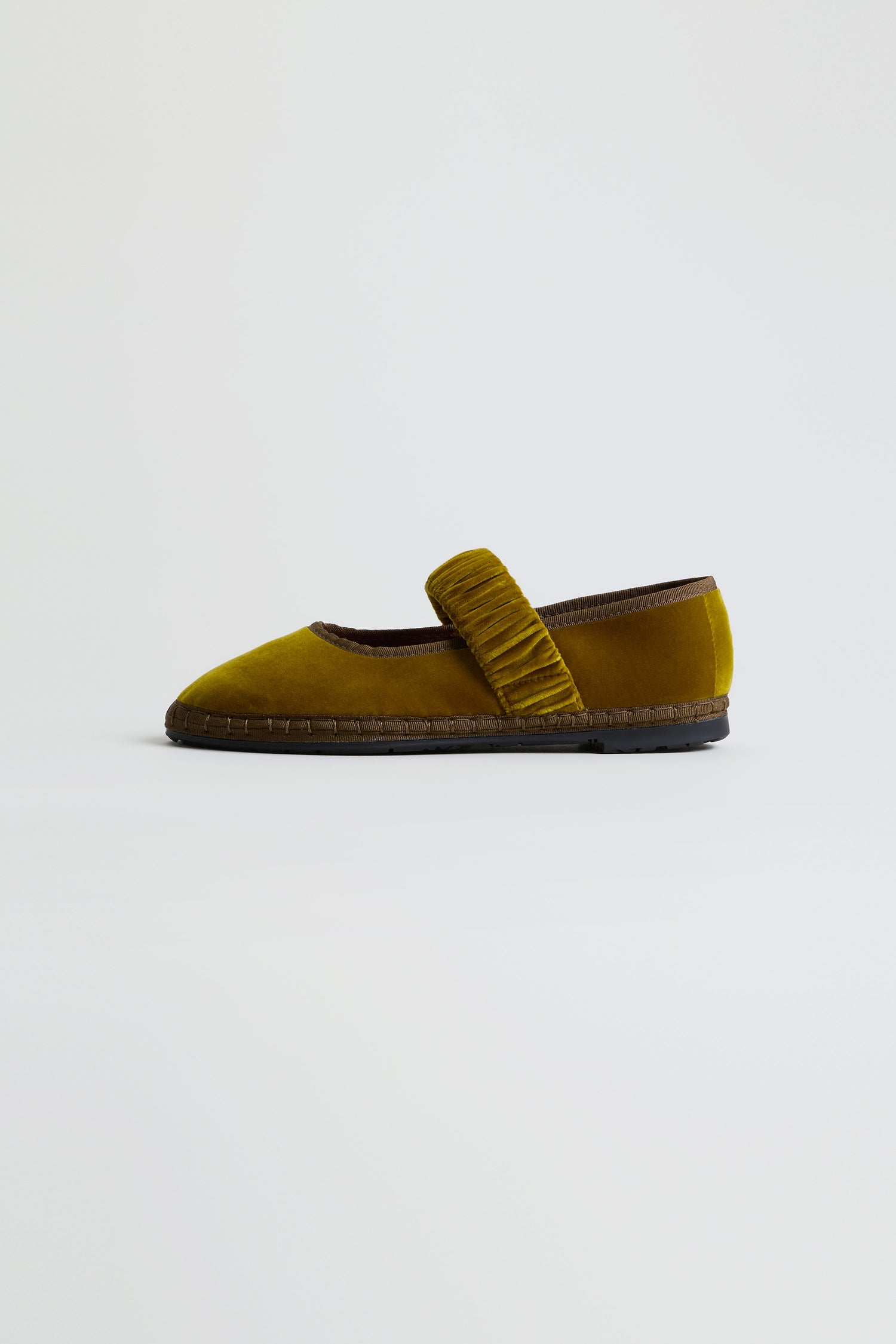 Introducing the Mafalda Ochre Velvet Shoes: a single mustard yellow velvet flat with a ruched strap across the top, complemented by brown trim and a flat sole crafted from recycled rubber. Handmade in Spain, this elegant velvet shoe is displayed against a plain light background.