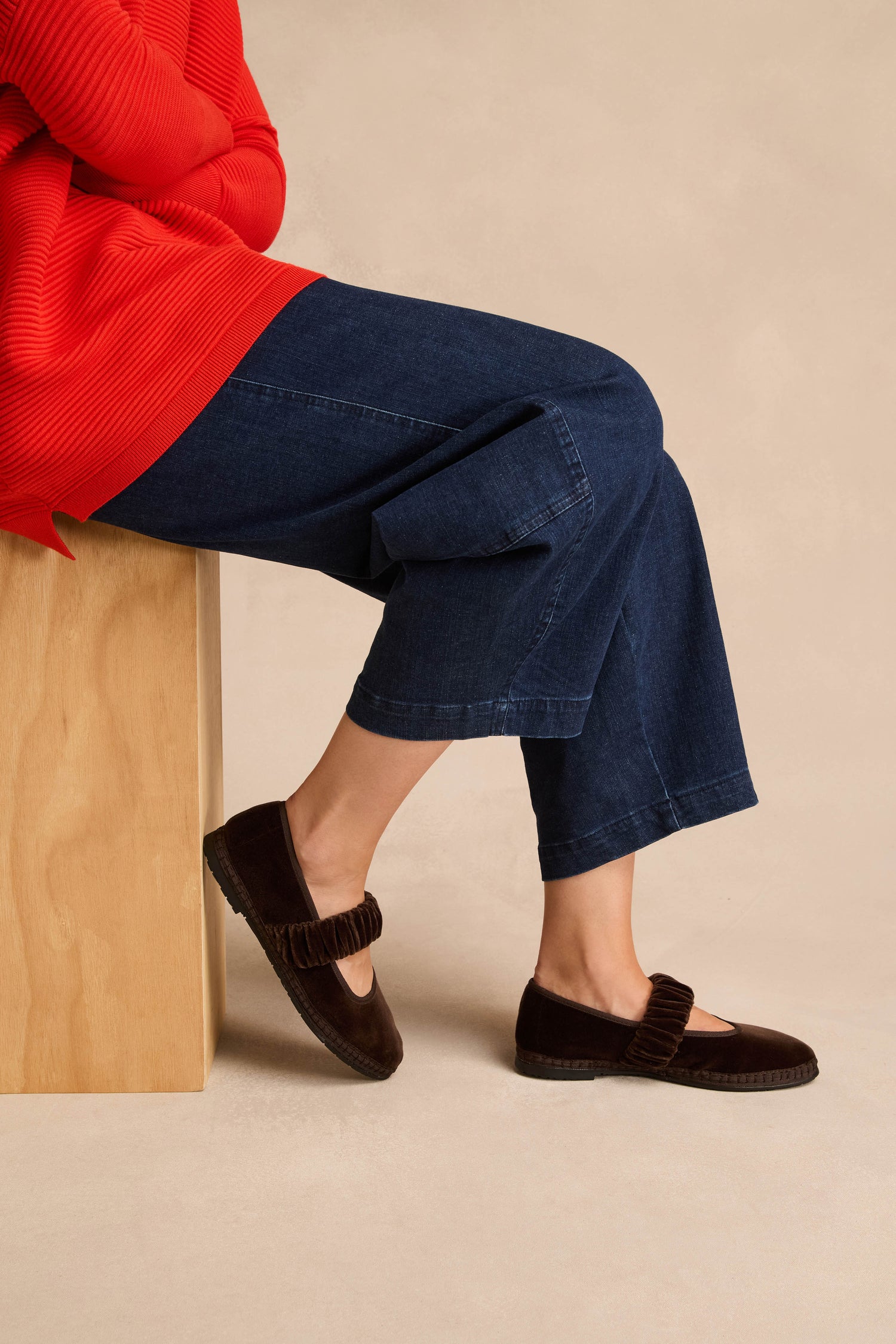 Wearing the Mafalda Truffle Velvet Shoes—featuring a ruffled strap detail and recycled rubber sole—a person in a red sweater and wide-leg blue jeans sits on a wooden bench.