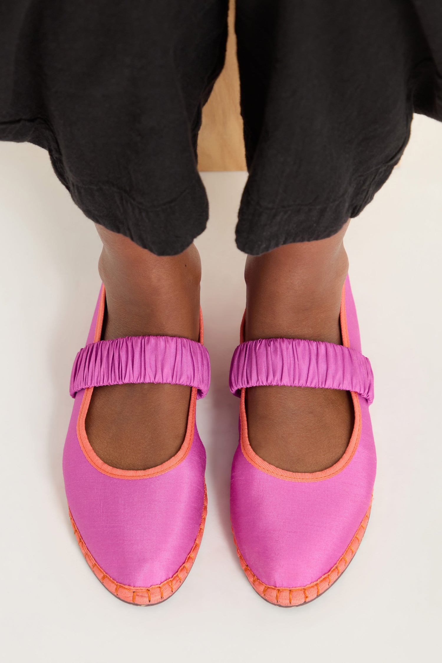 A person wearing black pants and Mafalda Silk Shoes—handcrafted in Spain in a bright pink Mary Jane style with orange trim—is standing on a light-colored floor.