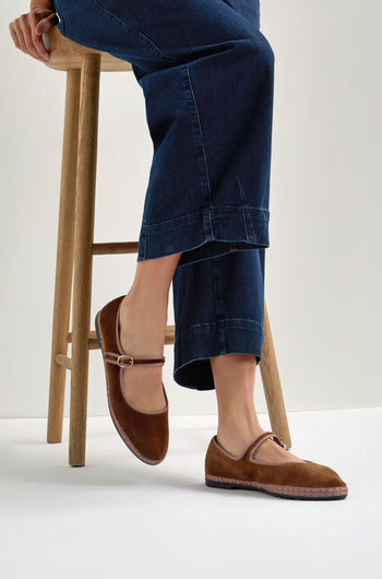 A person wearing dark blue jeans sits on a wooden stool, showcasing Brown Mary Jane Velvet Shoes featuring a small buckle that adds vintage charm to their classic look.