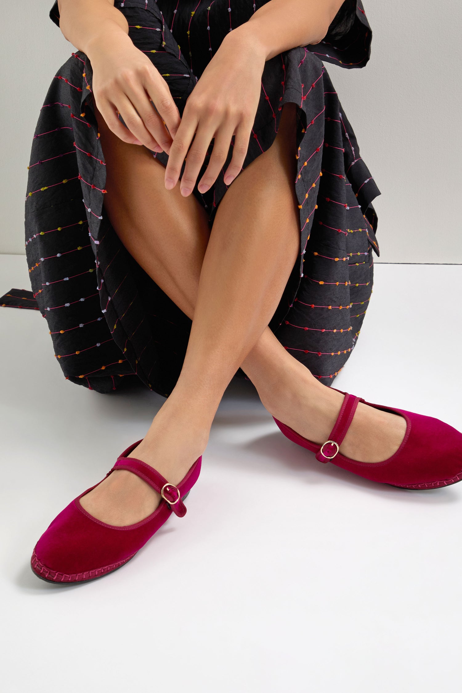 A person sits cross-legged, wearing a black dress with colorful stripes and vintage charm, paired with fuchsia Mary Jane velvet shoes.