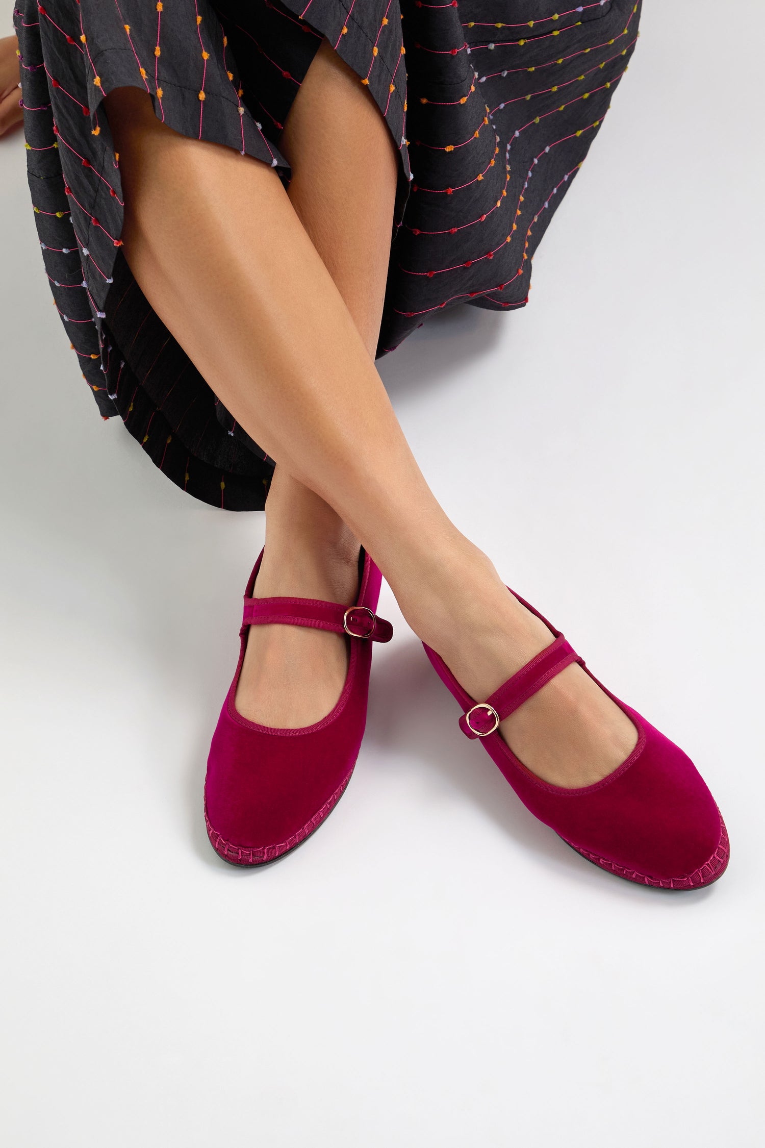 A person in a dark skirt with colorful stitching sits cross-legged, showcasing Fuchsia Mary Jane Velvet Shoes with vintage-style buckles.