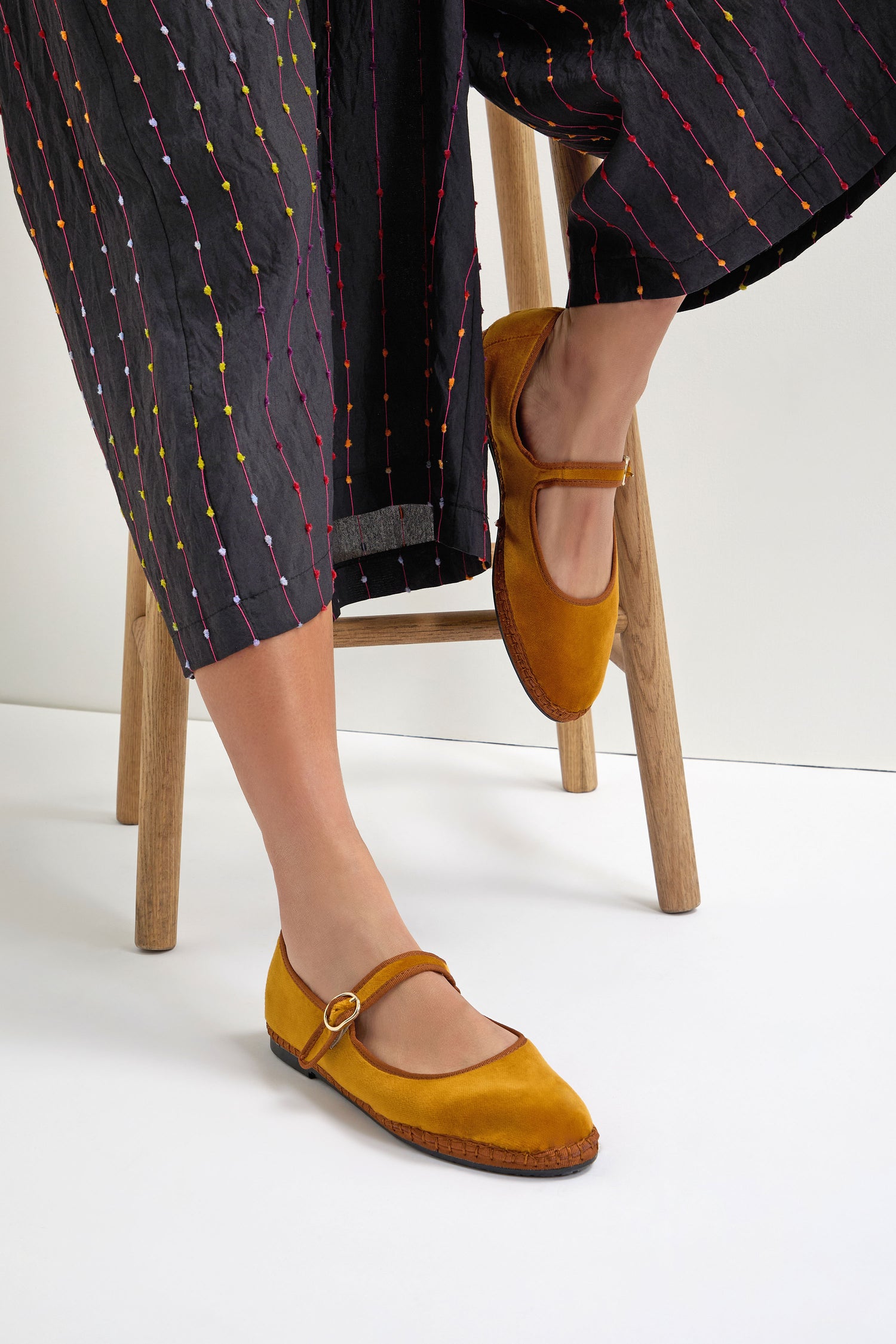 Seated on a wooden stool, the person wears Mustard Mary Jane Velvet Shoes—crafted with sustainable Spanish shoemaking—featuring a round buckle, paired with black pants with colorful vertical stripes.