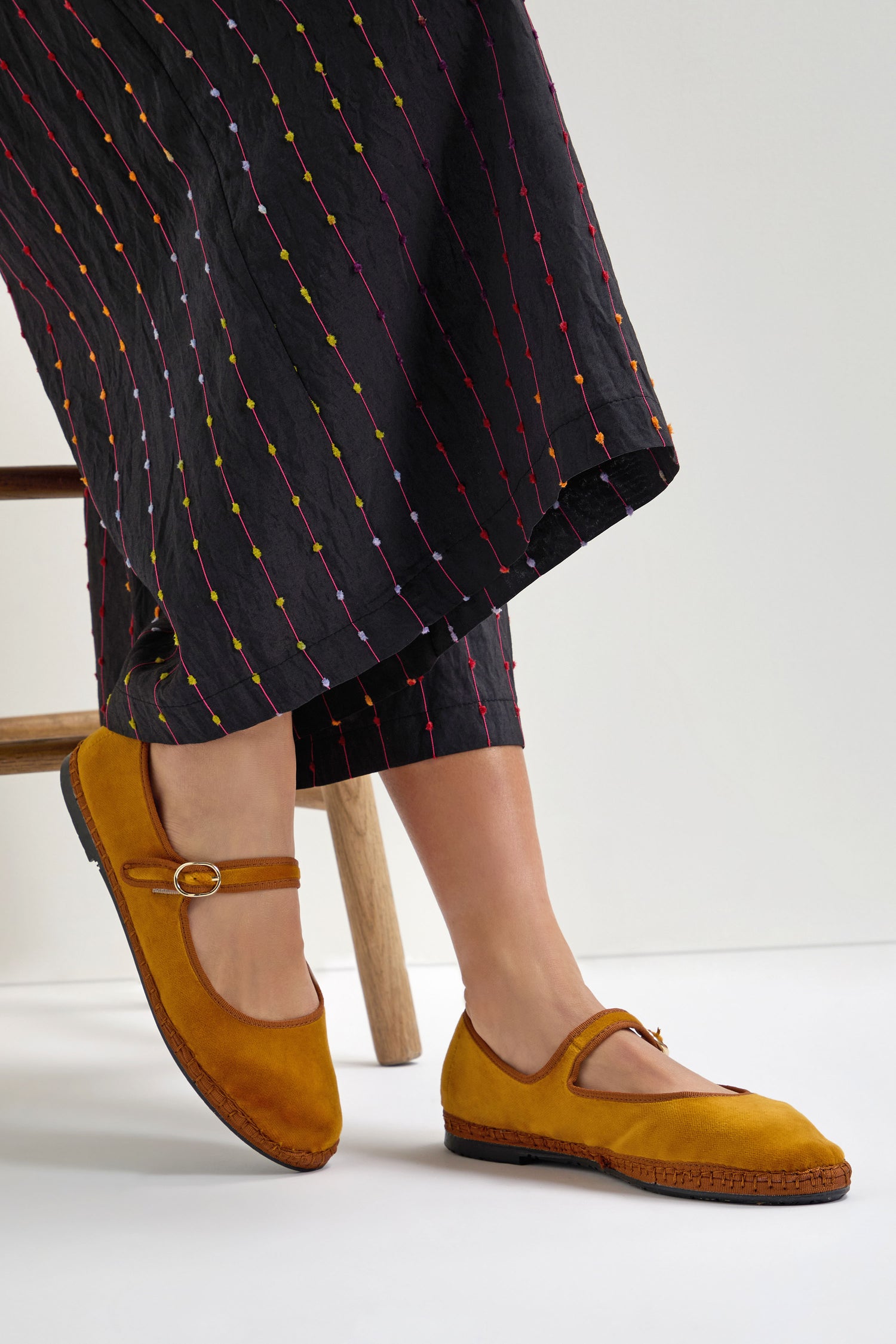 A person wearing Mustard Mary Jane Velvet Shoes, crafted sustainably, and black pants with colorful vertical stitching sits on a wooden chair.