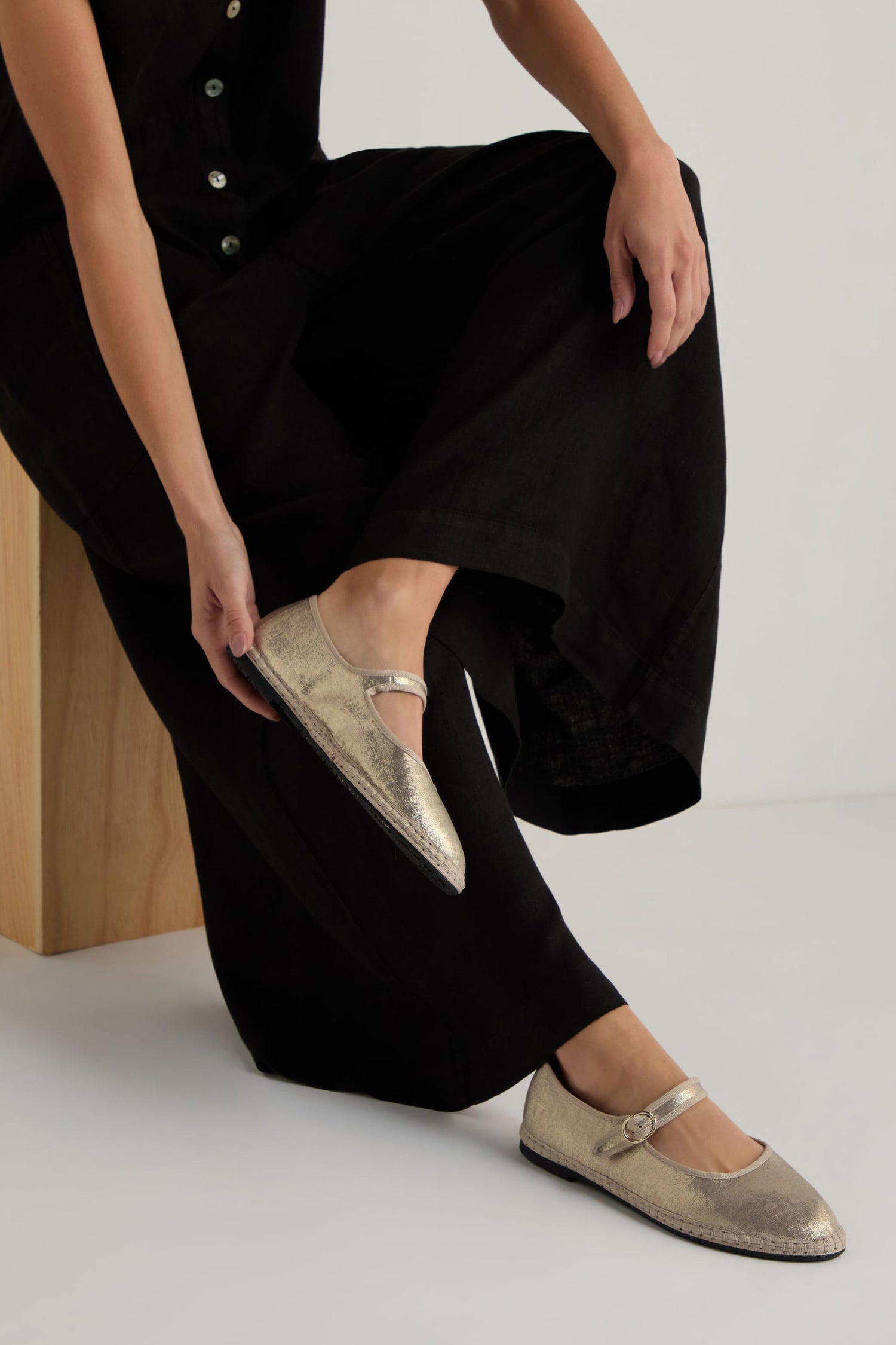 Someone in black pants sits on a wooden bench, adjusting their Metallic Mary Jane Shoes, which feature a recycled bicycle tyre sole.