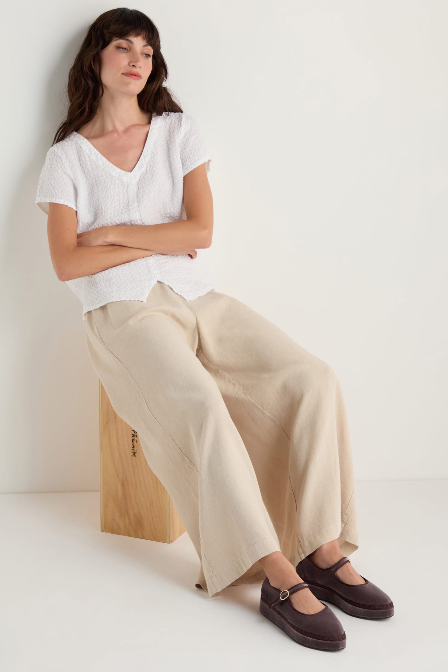 Woman with long brown hair sits on a wooden block, wearing a white short-sleeve top, wide-leg beige pants, and Taupe Velvet Platform Mary Jane Shoes with a cotton velvet upper, handmade in Spain, against a light background.