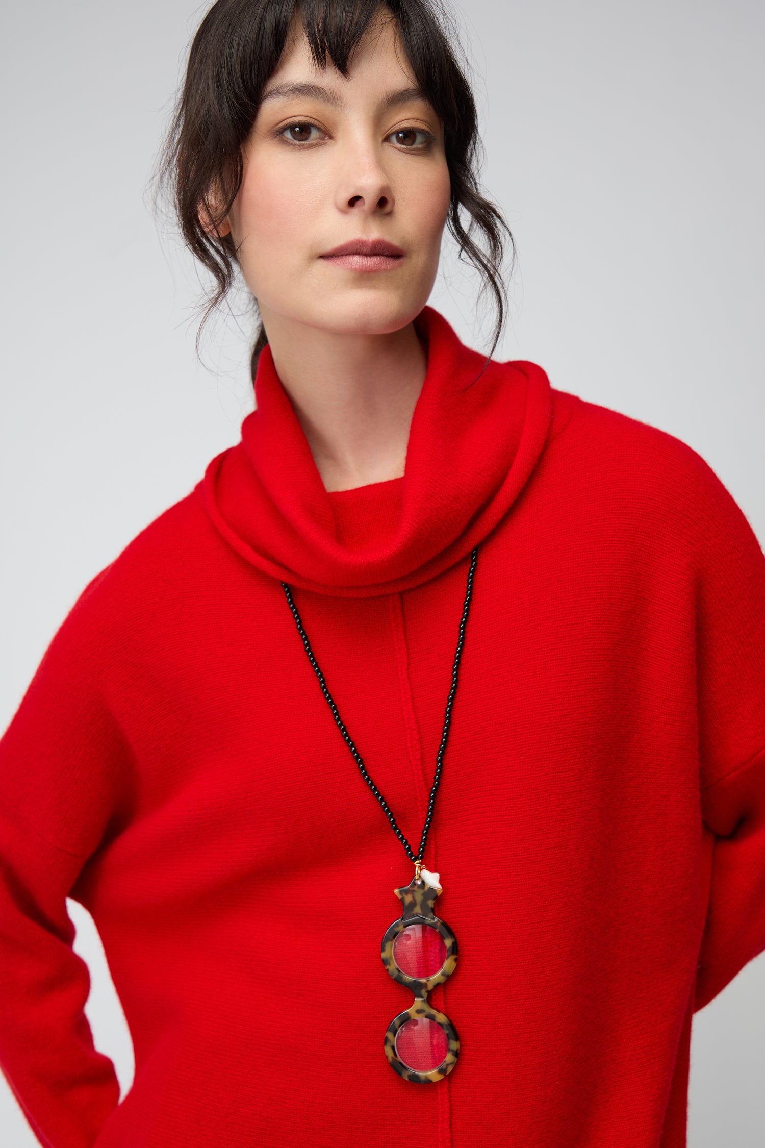 Woman wearing a bright red cowl-neck sweater and a long Star Pendant Glasses necklace, featuring a magnifying lens pendant, standing against a neutral background.