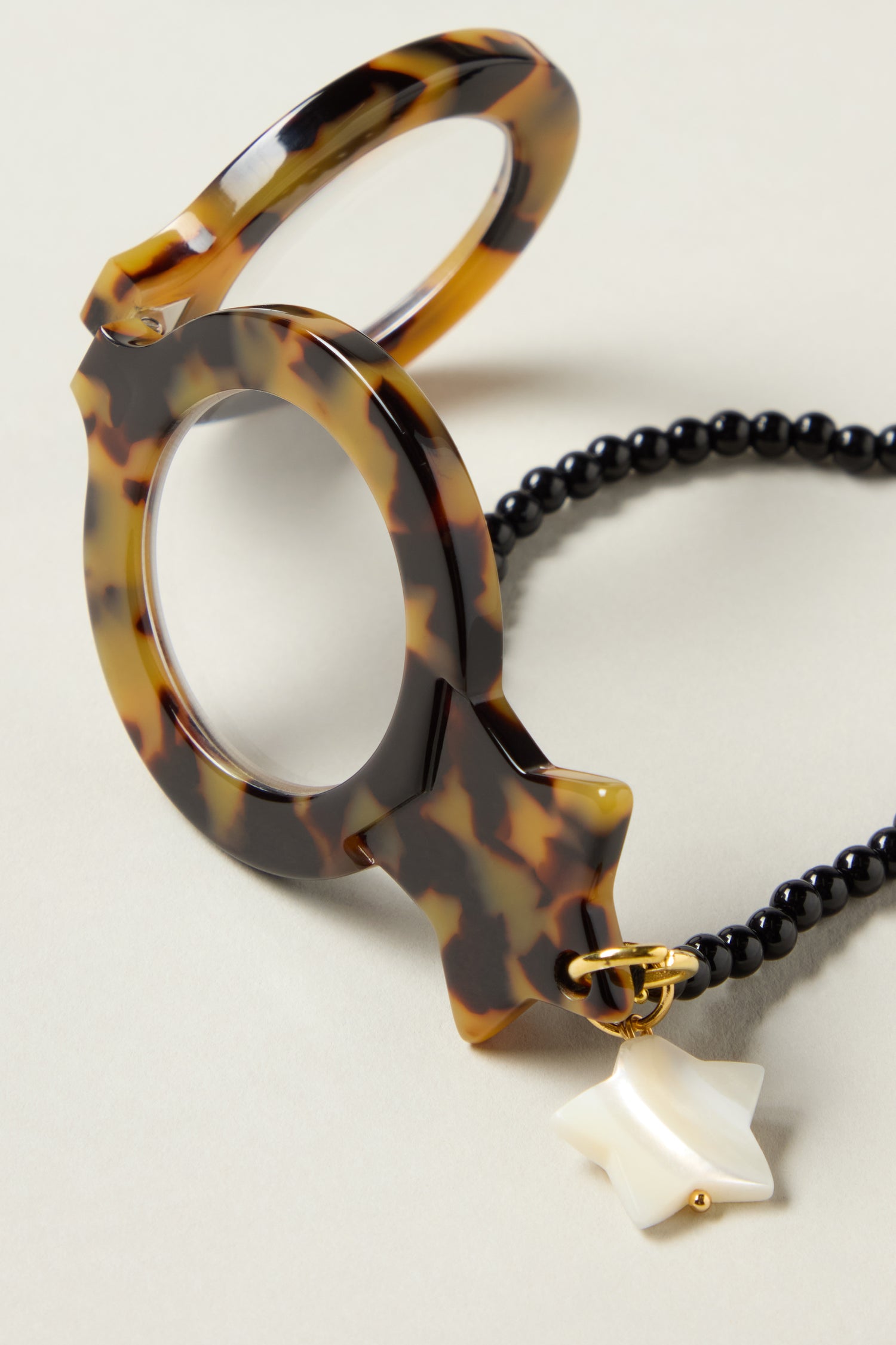 The Star Pendant Glasses are handcrafted tortoiseshell eyewear with a gold star charm and black beaded chain, featuring magnifying lens pendants, displayed elegantly on a neutral surface.