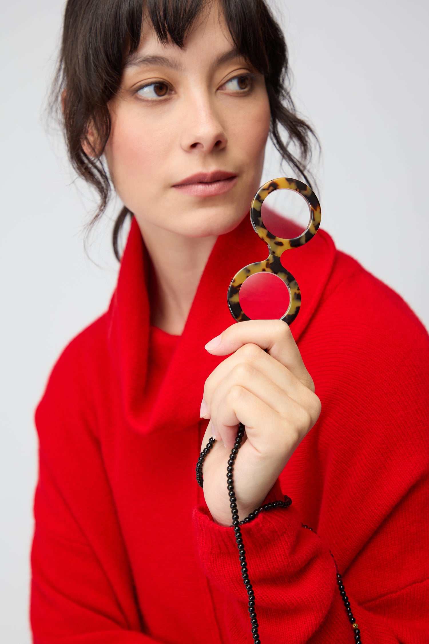 A woman in a red sweater holds Star Pendant Glasses—round, tortoiseshell frames with a black chain—and gazes to the side against a plain background.