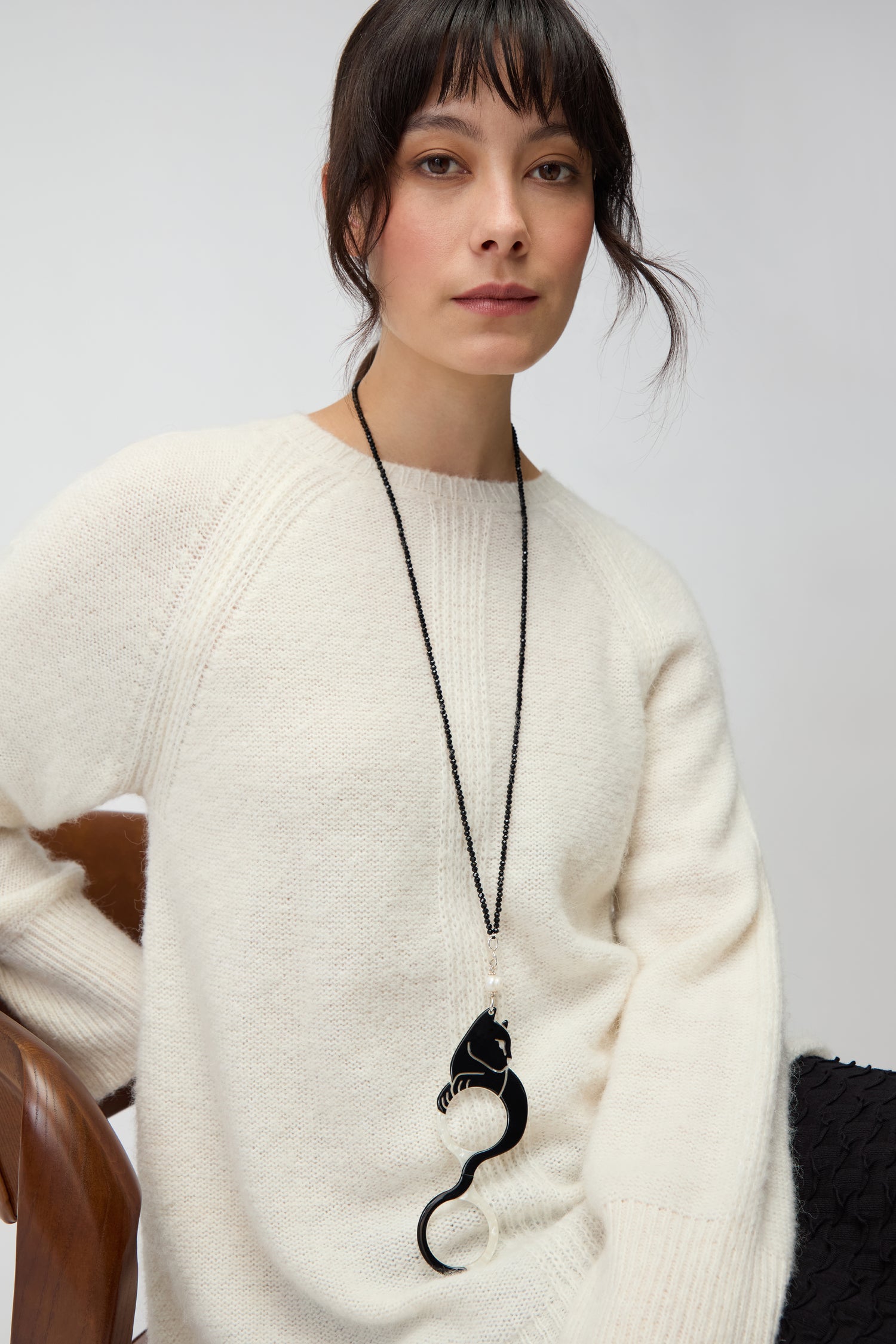 A woman seated on a wooden chair wears an off-white knit sweater and the Cat Pendant Glasses—a long black necklace featuring a large, abstract magnifying pendant—against a plain background.