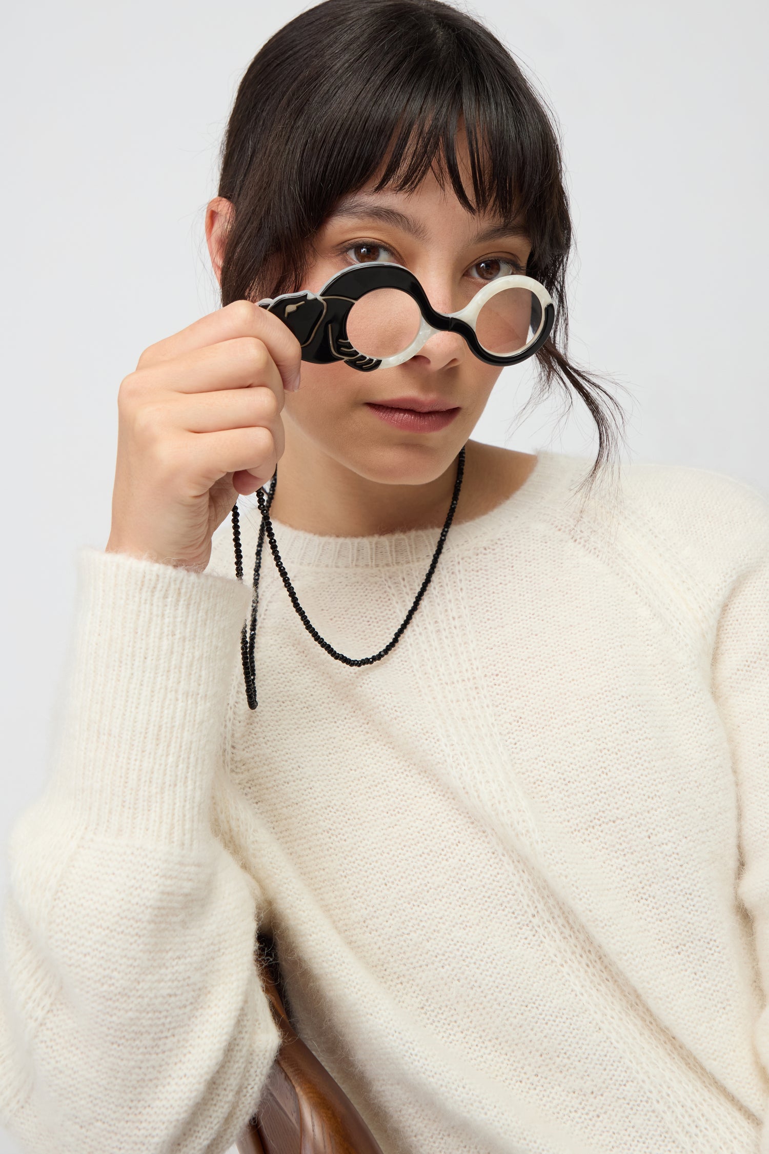 A woman with dark hair and bangs wears a cream sweater and holds Cat Pendant Glasses, a pair of round black eyeglasses, raising one lens in front of her face.