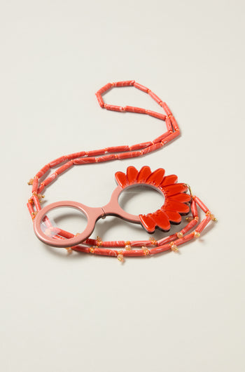 The Daisy Pendant Glasses feature a coral-hued beaded eyeglass chain with a daisy pendant holder, elegantly displayed on a neutral surface.
