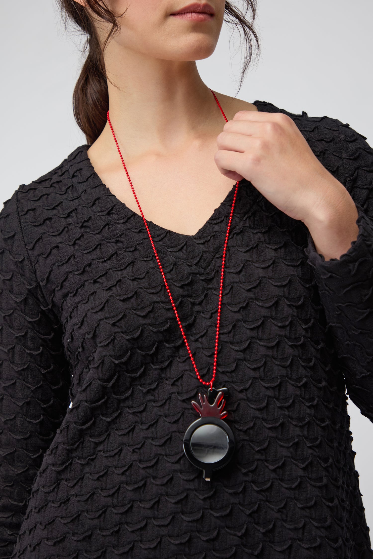 A woman wears a textured black top and the Revolver Pendant Glasses—a long red necklace with a large circular black optical-grade pendant featuring a flame-shaped detail.