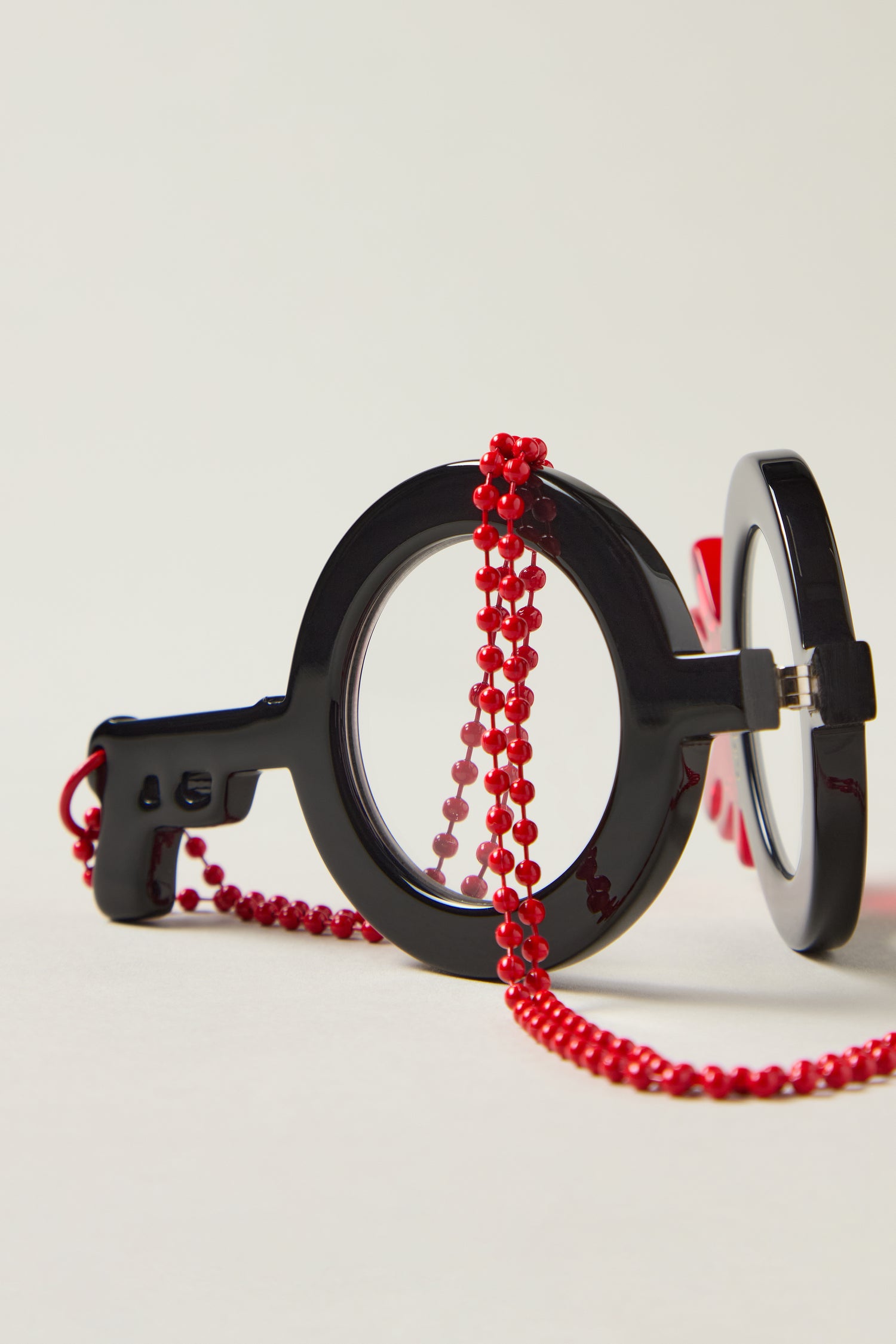Revolver Pendant Glasses are black, key-shaped frames with clear lenses, adorned with a red beaded chain—an eye-catching piece of optical-grade jewelry displayed against a plain background.