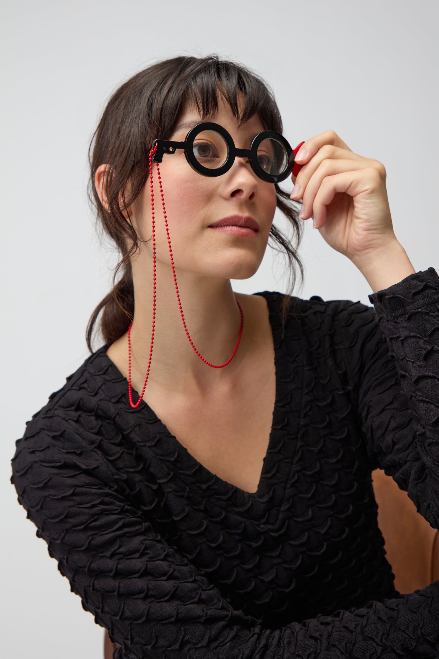 A woman in a textured black top holds up Revolver Pendant Glasses, featuring a red beaded chain—showcasing quirky style and optical-grade jewellery flair.