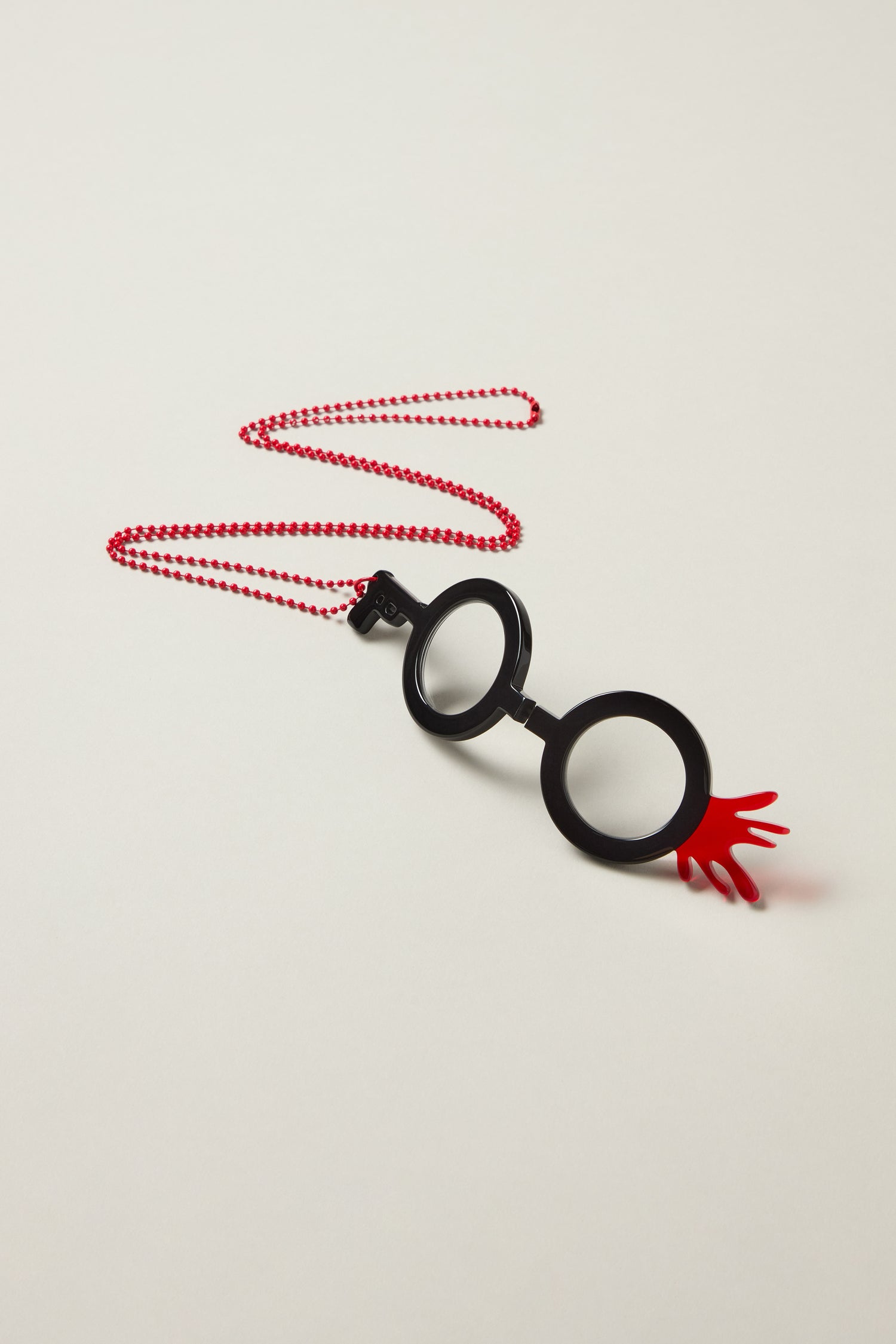 The Revolver Pendant Glasses is a necklace with a red chain and a black, eyeglass-shaped circular pendant, crafted as optical-grade jewellery, featuring a small red hand detail on the lower right.