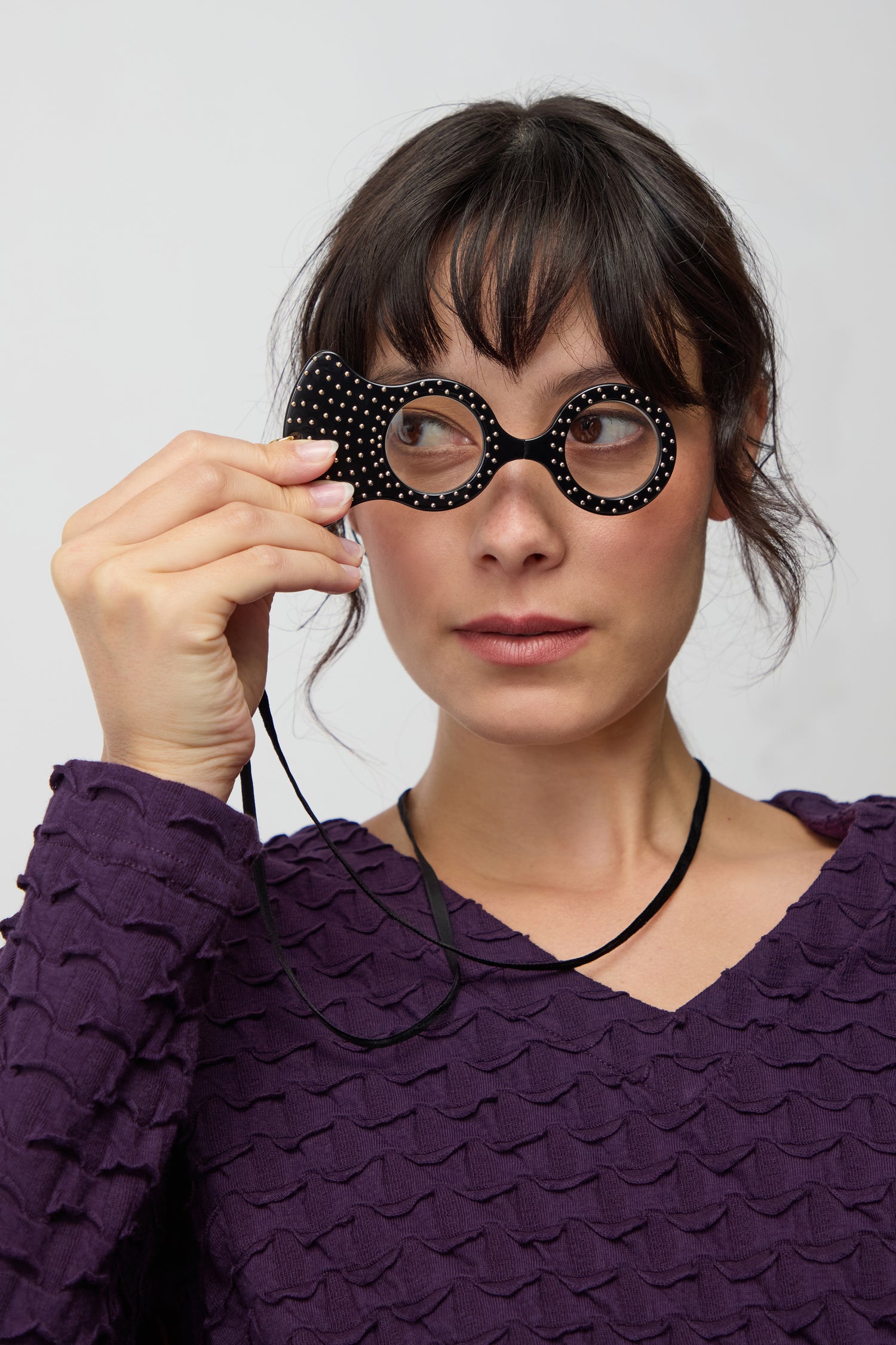 A woman in a purple textured top holds up the Rock and Roll Pendant Glasses, a decorative black magnifying lens, to her right eye while looking to the side.