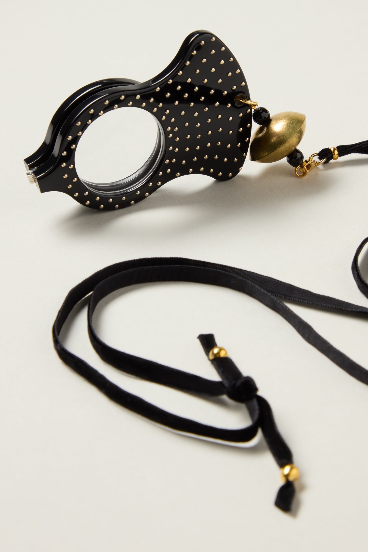 The Rock and Roll Pendant Glasses feature a black magnifying glass pendant with gold studs and a large gold bead, handmade in Spain, attached to a black velvet ribbon with gold accents and displayed on a light surface.