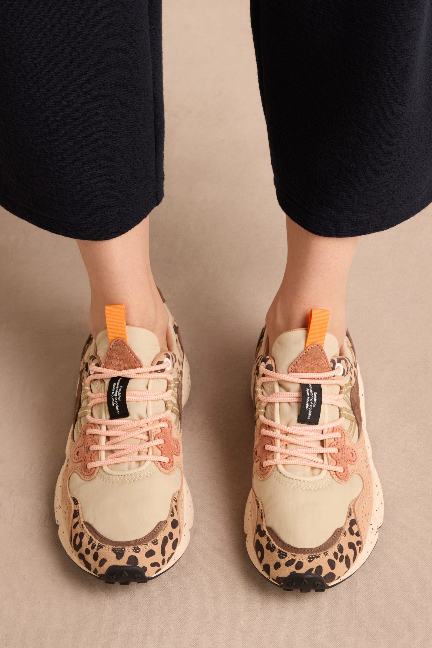 A person wearing beige pants and cream-colored Yamano 3 Cheetah Detail Trainers with suede, cheetah print accents, pink laces, and orange tabs stands on a light surface.