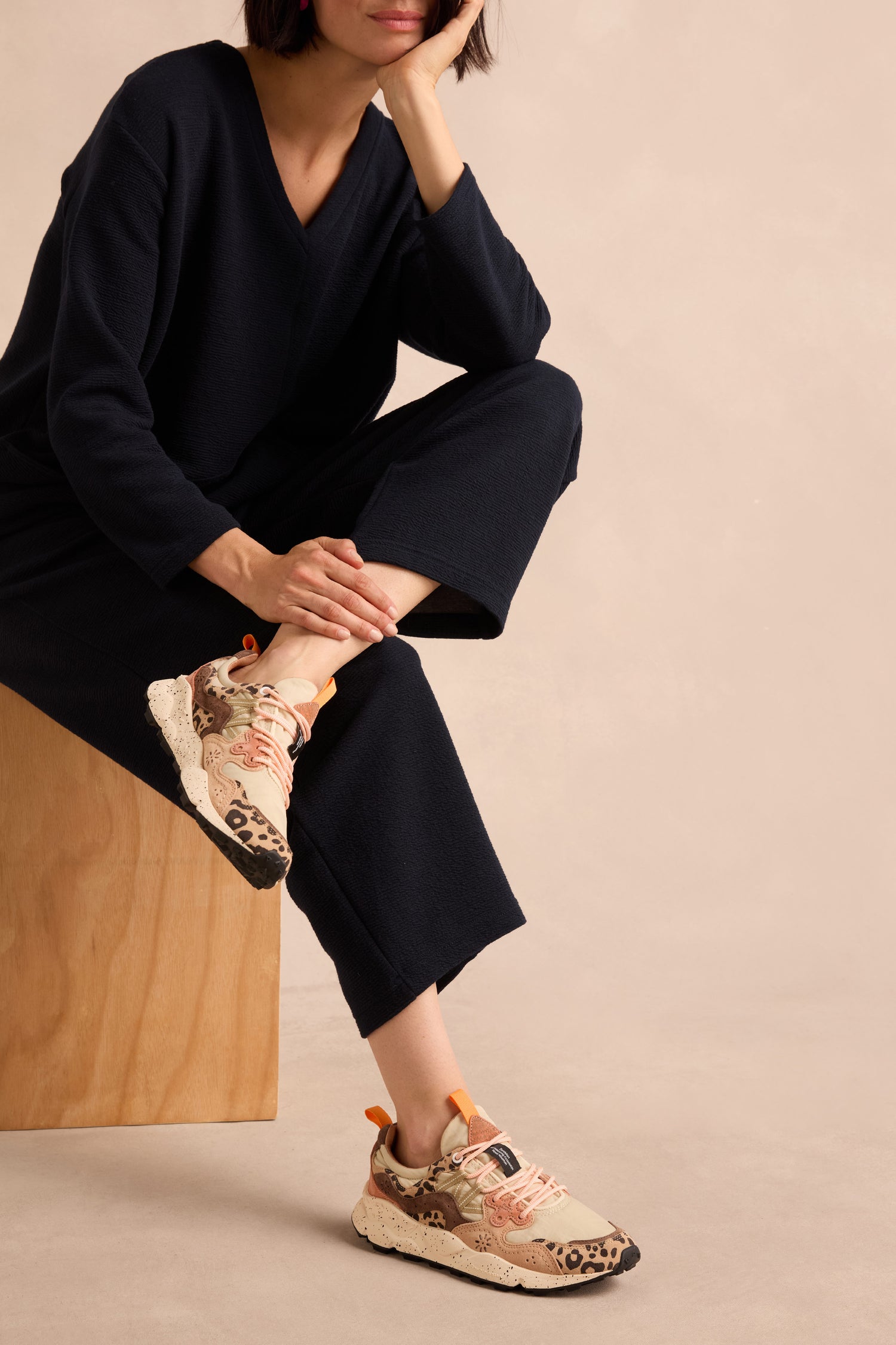 A person seated on a light wooden bench against a neutral background wears beige pants and Yamano 3 Cheetah Detail Trainers featuring suede uppers, pink laces, and cheetah-patterned accents.