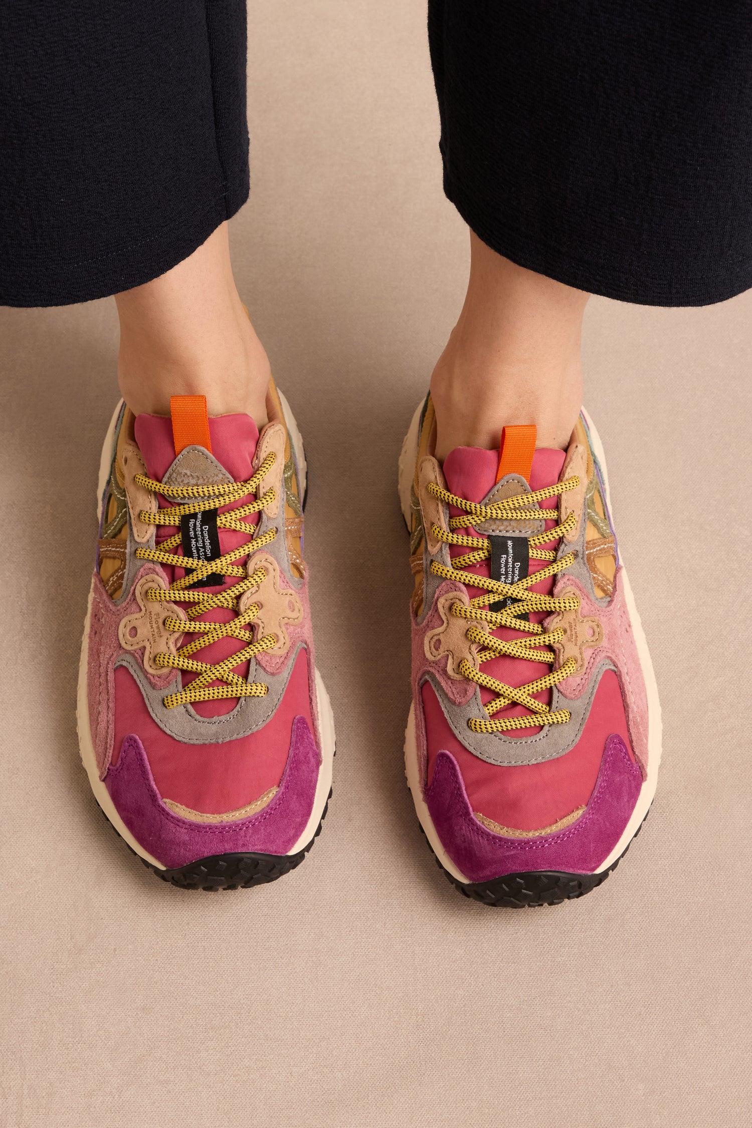Top view of a person wearing light-colored pants and the Solin Colour Pop Trainers by Flower Mountain, crafted from suede and technical fabric with pink, purple, and yellow accents and multicolored laces—these vibrant trainers stand out beautifully against the beige surface.