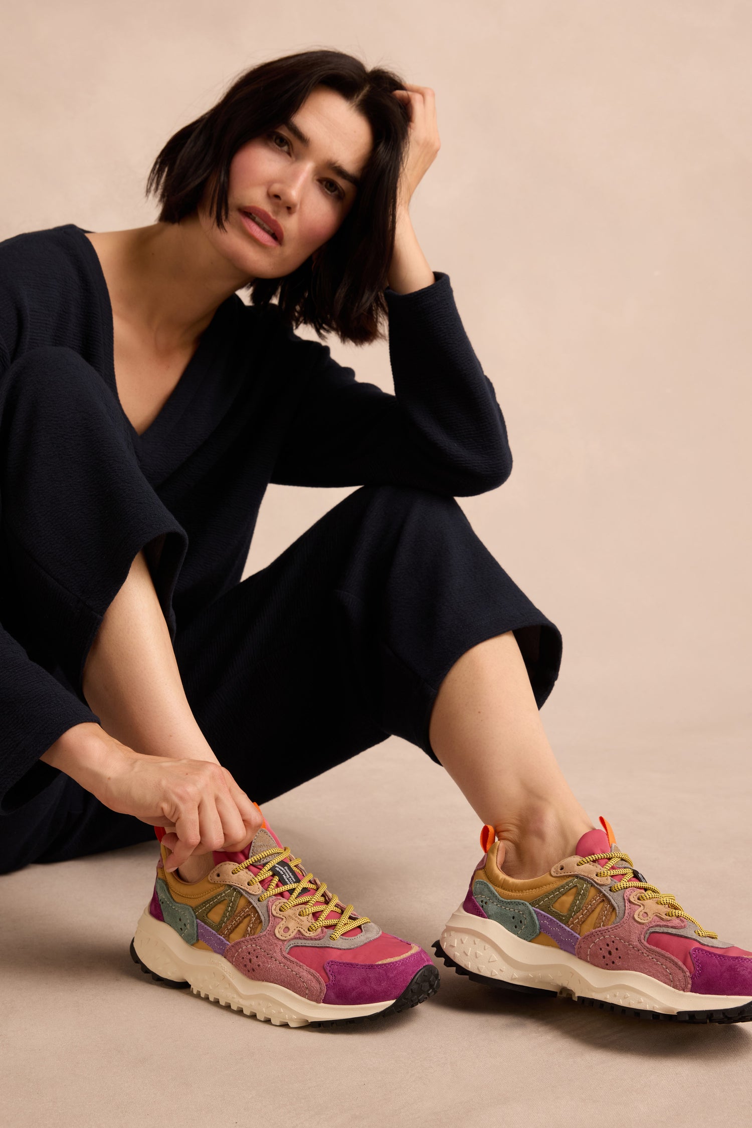 A person wearing beige pants and Solin Colour Pop trainers—crafted from suede and technical fabric with vibrant purple, green, tan, and orange accents—seated on a wooden chair against a neutral background.