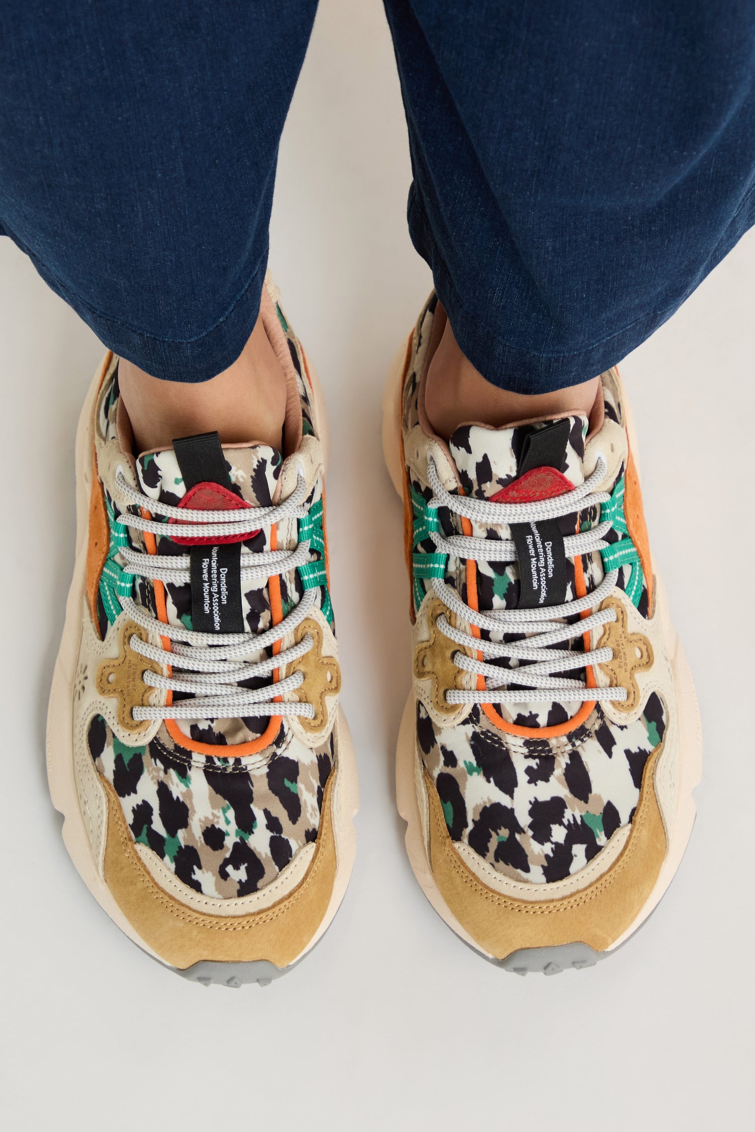 A person wearing Yamano 3 Leopard Suede Nylon Trainers with colorful leopard print, green accents, beige suede details, and a lightweight rubber sole, paired with dark blue pants on a light surface.