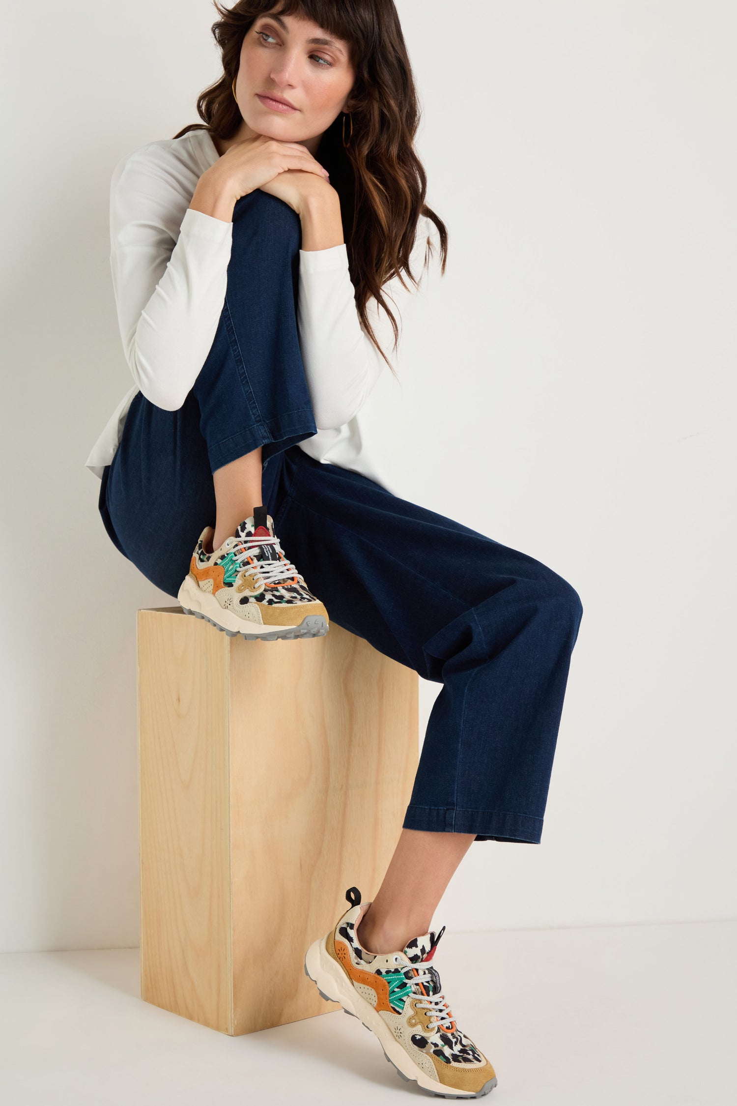 A woman in a white long-sleeve shirt and blue jeans sits on a wooden box with her arms on her knees, wearing Yamano 3 Leopard Suede Nylon Trainers featuring a lightweight rubber sole.