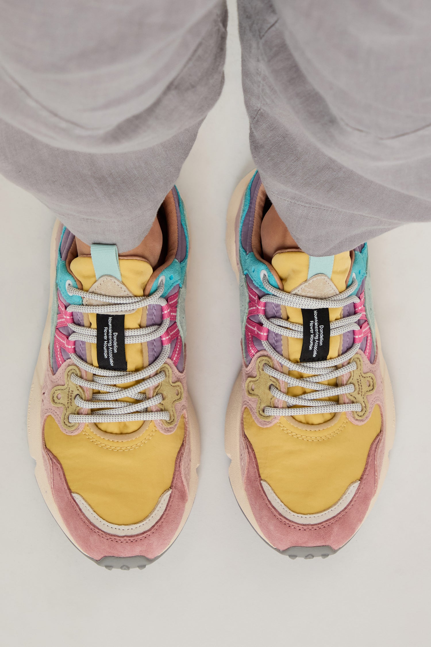 A person wearing light gray pants and Dijon Yamano 3 Colour Pop Trainers with yellow, pink, blue, and beige panels, viewed from above.