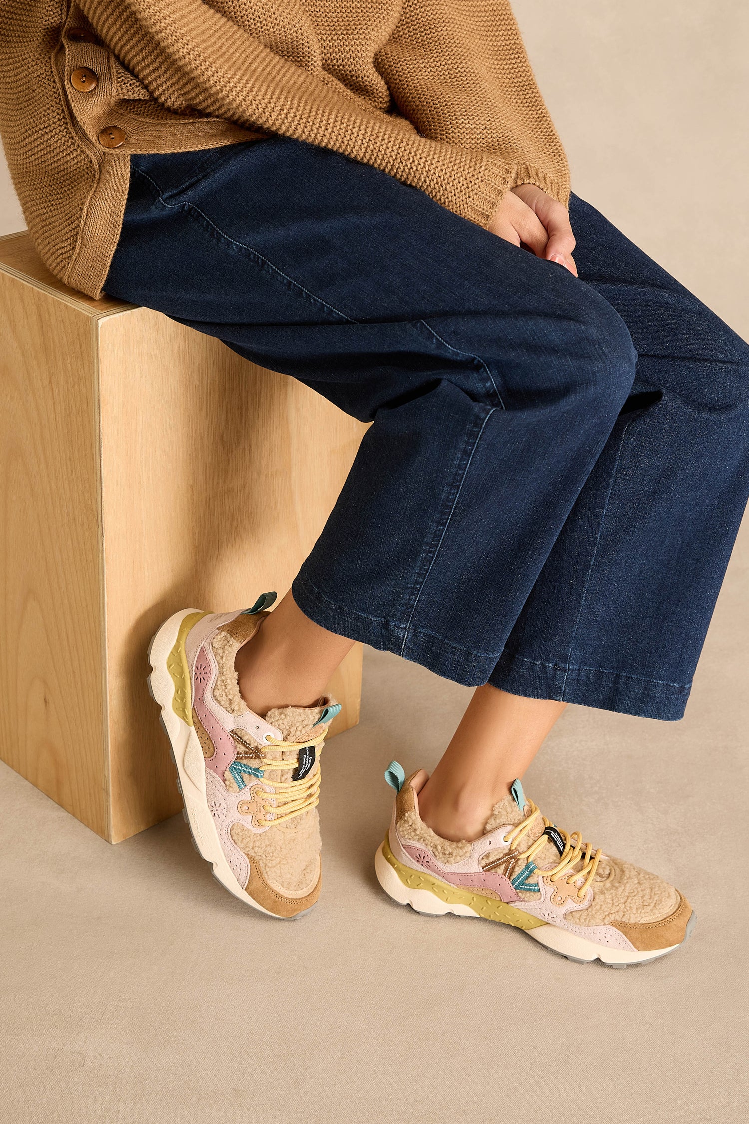 A person wearing wide-leg jeans, a brown sweater, and Yamano 3 Teddy Trainers featuring a comfy cork insole sits on a wooden block against a beige background.