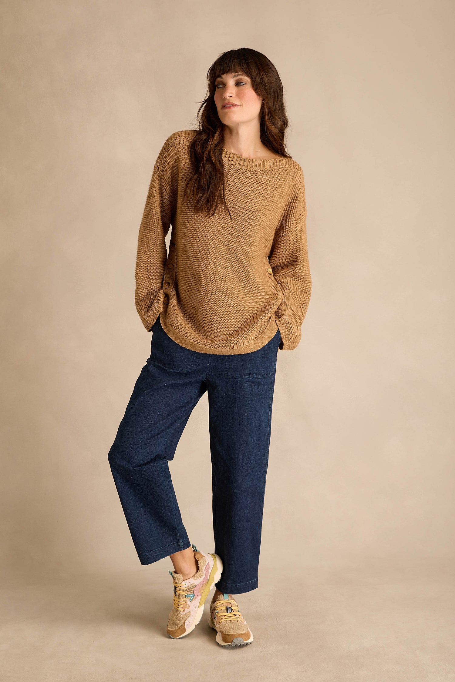 A woman stands against a beige background with her hands in her pockets, wearing a tan sweater, blue wide-leg pants, and Yamano 3 Teddy Trainers—tan sneakers featuring cork insoles.