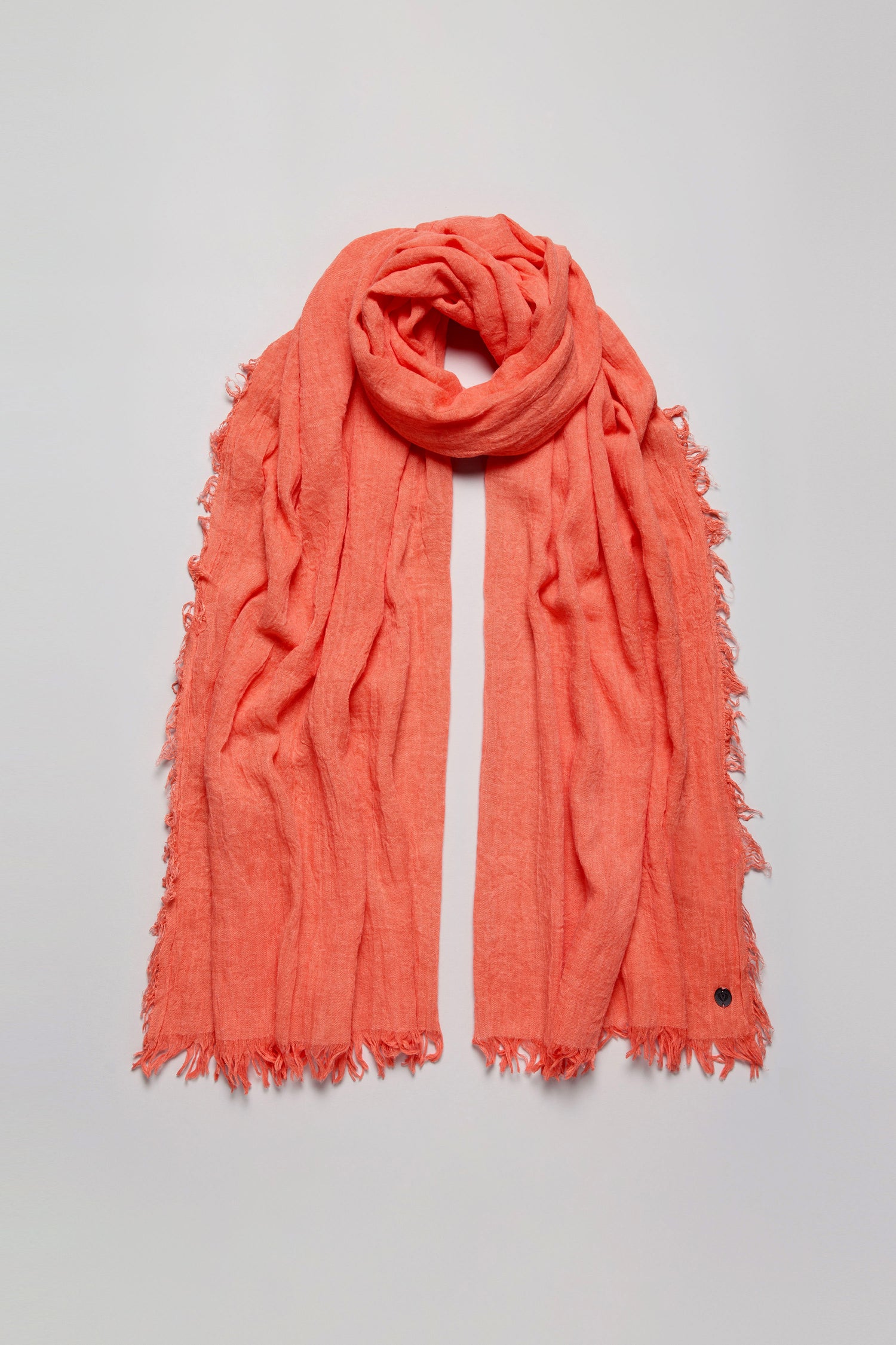 The Soft Fringe Scarf in bright coral features frayed edges and textured material, shown laid flat on a light grey surface. Ideal for versatile styling year-round.