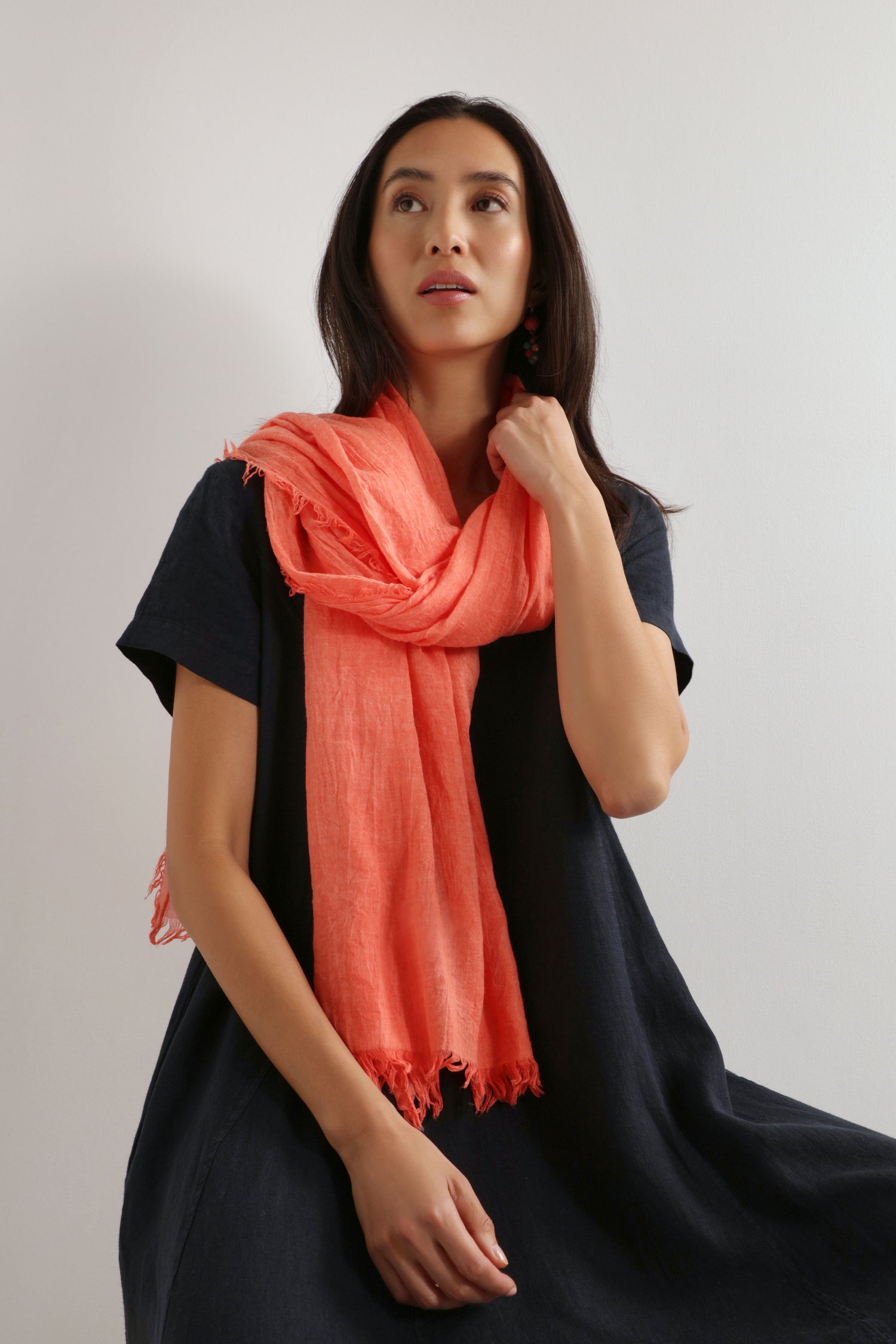 A woman with long dark hair models the Soft Fringe Scarf in coral over a loose black dress, sitting against a plain light background and gazing upward, highlighting the scarf’s versatile style.