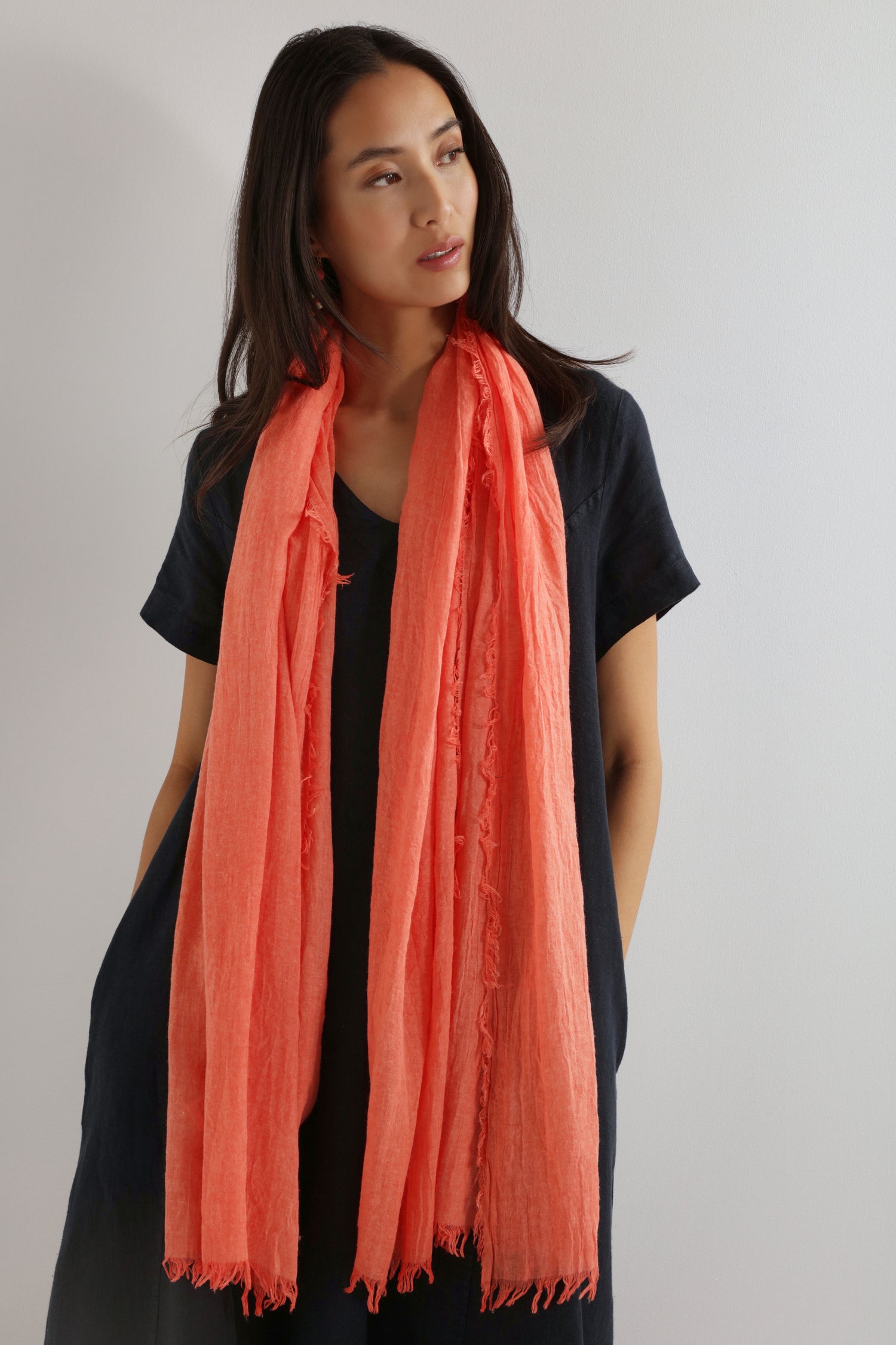 A woman in a black dress models the Soft Fringe Scarf in orange, featuring textured material and fringe detail, standing against a plain light background and looking to the side.