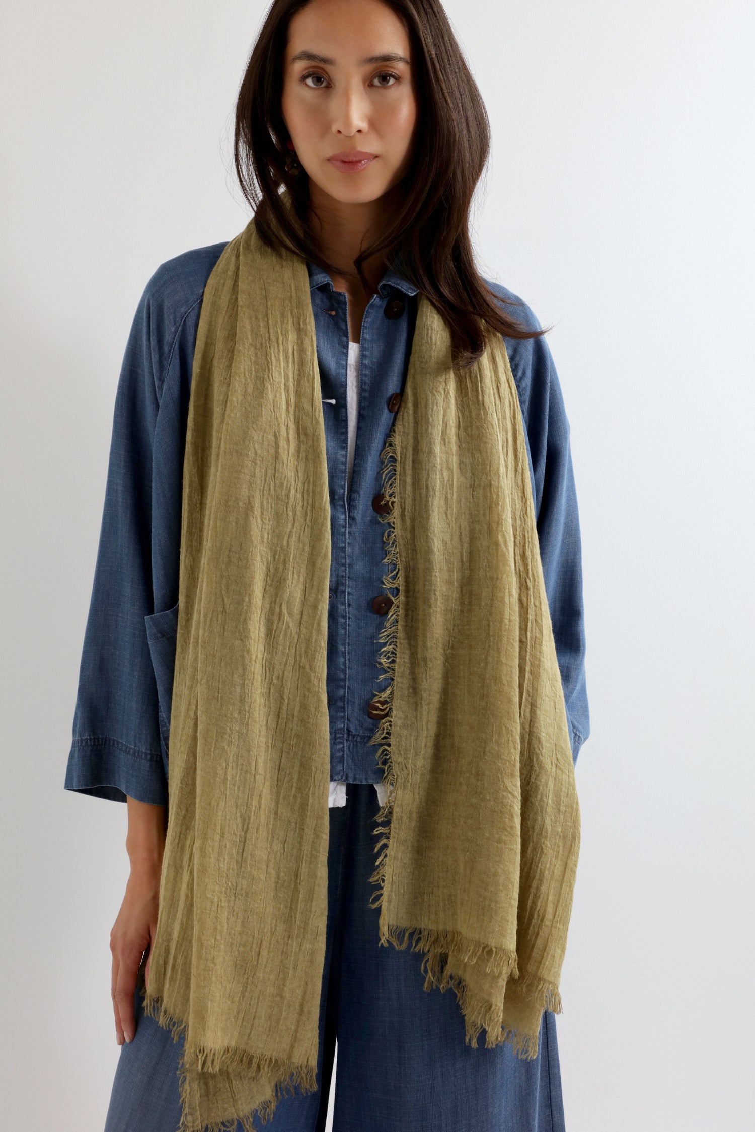 A woman in a blue button-up shirt and wide-leg pants stands against a plain background, wearing the Soft Fringe Scarf, which features textured material and fringe edges—ideal for versatile styling.