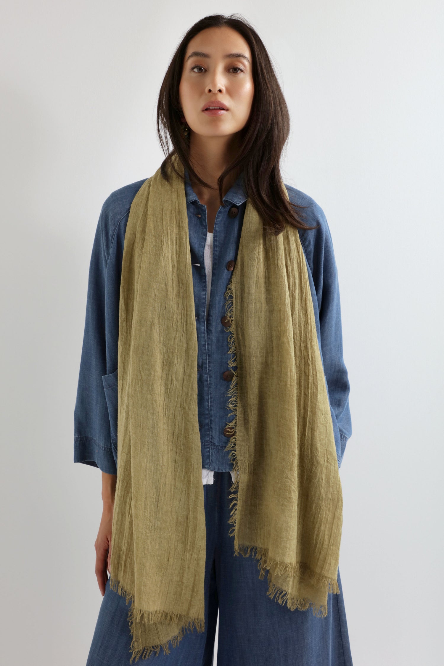 A woman stands against a plain background in a blue denim shirt and pants, wearing the Soft Fringe Scarf in light olive-green textured material draped around her neck for versatile styling.