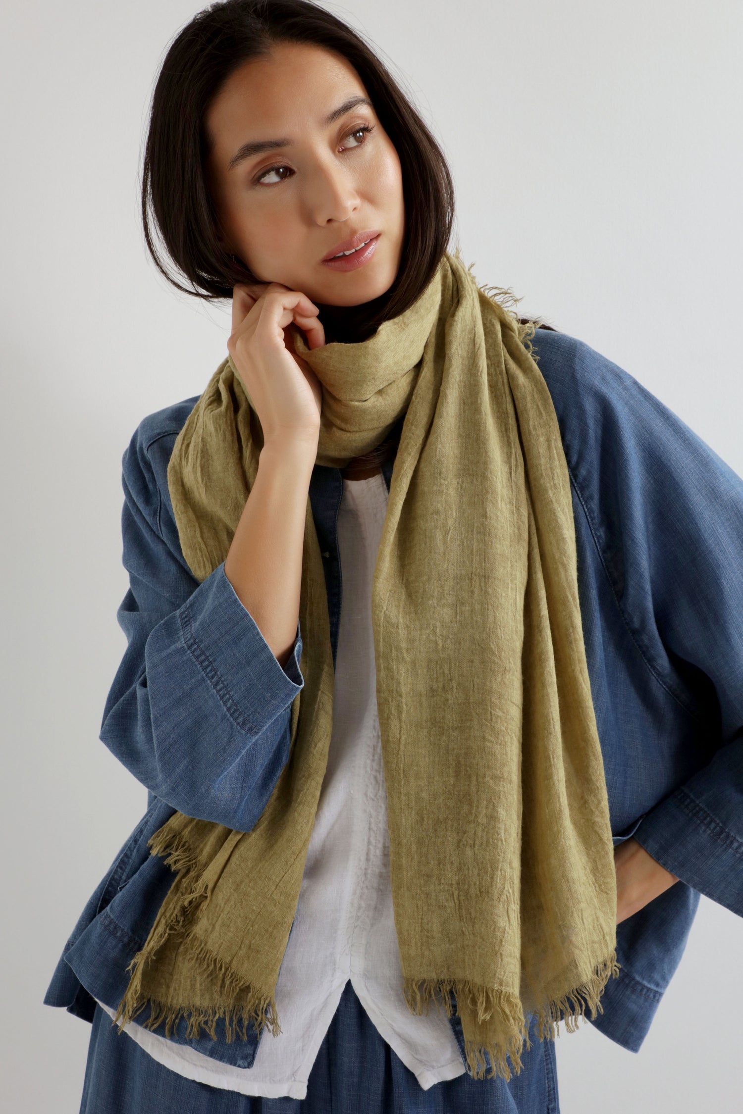 A woman wears the Soft Fringe Scarf in light olive over a blue denim jacket and white top, touching her neck as she looks to the side, highlighting the scarf’s versatile styling and textured material.