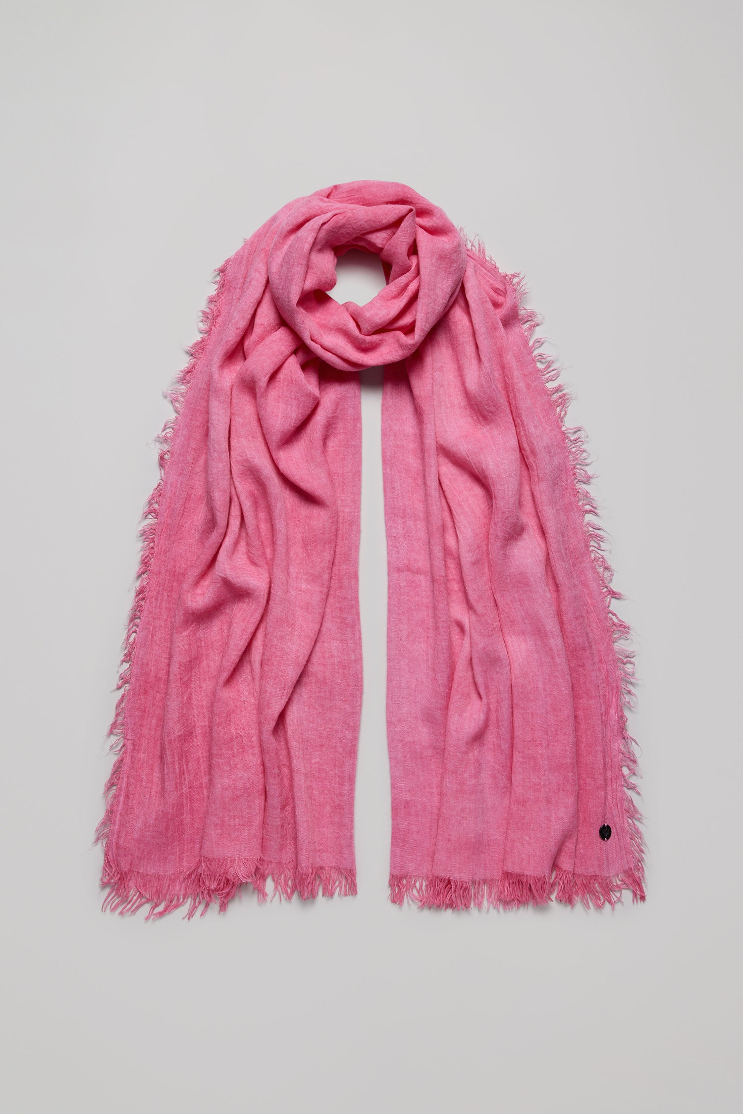 A Soft Fringe Scarf is displayed flat on a light background, highlighting its pink hue, frayed edges, and textured fabric for versatile styling.