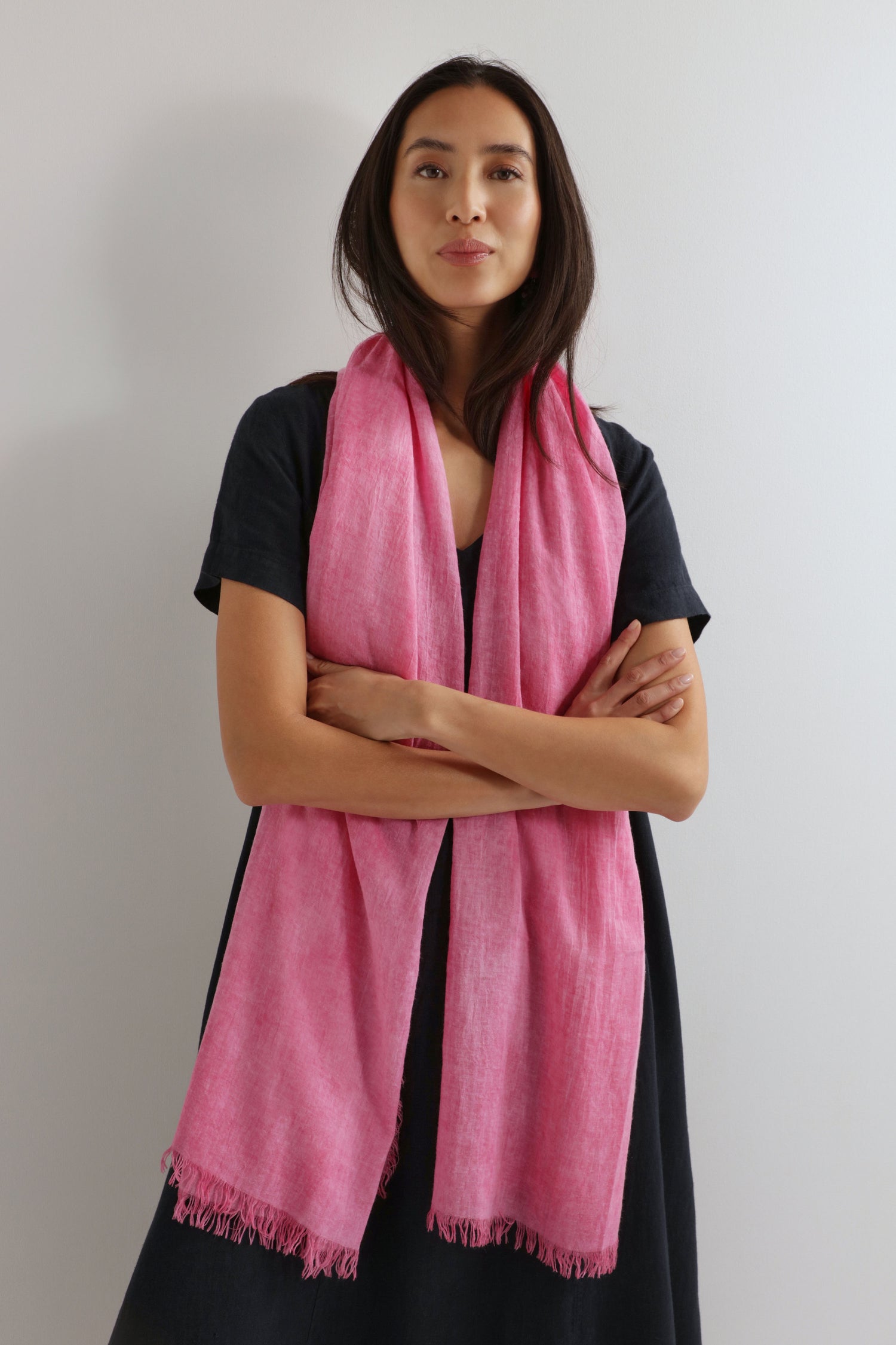 Woman stands against a plain wall, dressed in black and wearing the Soft Fringe Scarf—pink with textured material and fringed edges—arms crossed. This scarf is perfect for versatile styling.